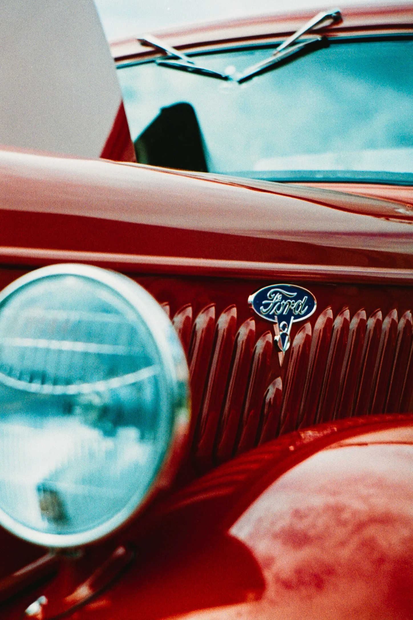 Close-up of a vintage red Ford car, focusing on the front grille with the Ford emblem and a large round headlamp in the foreground.