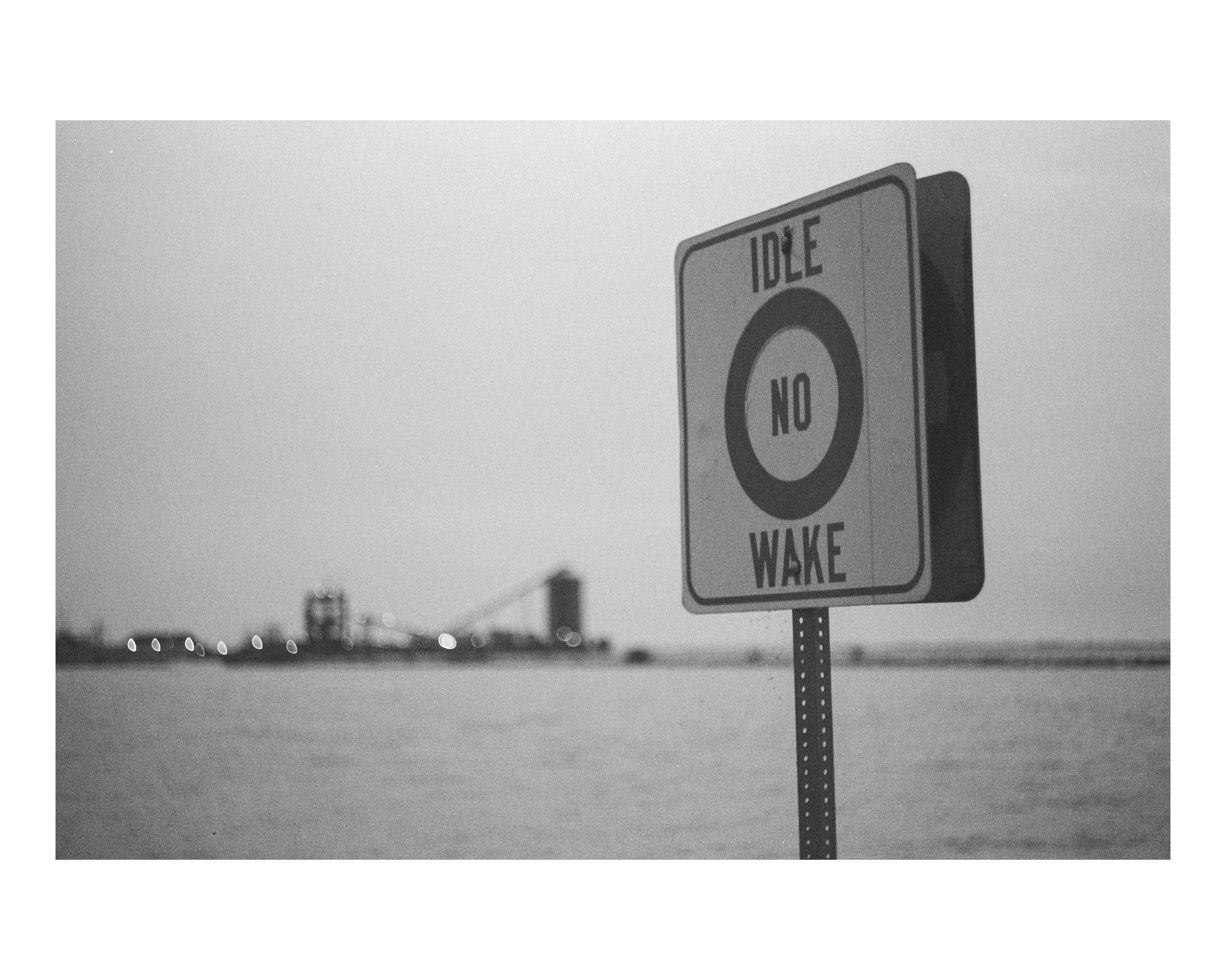 A black and white photo of a sign that reads 'Idle No Wake' near a body of water with a city skyline and bridge in the background.