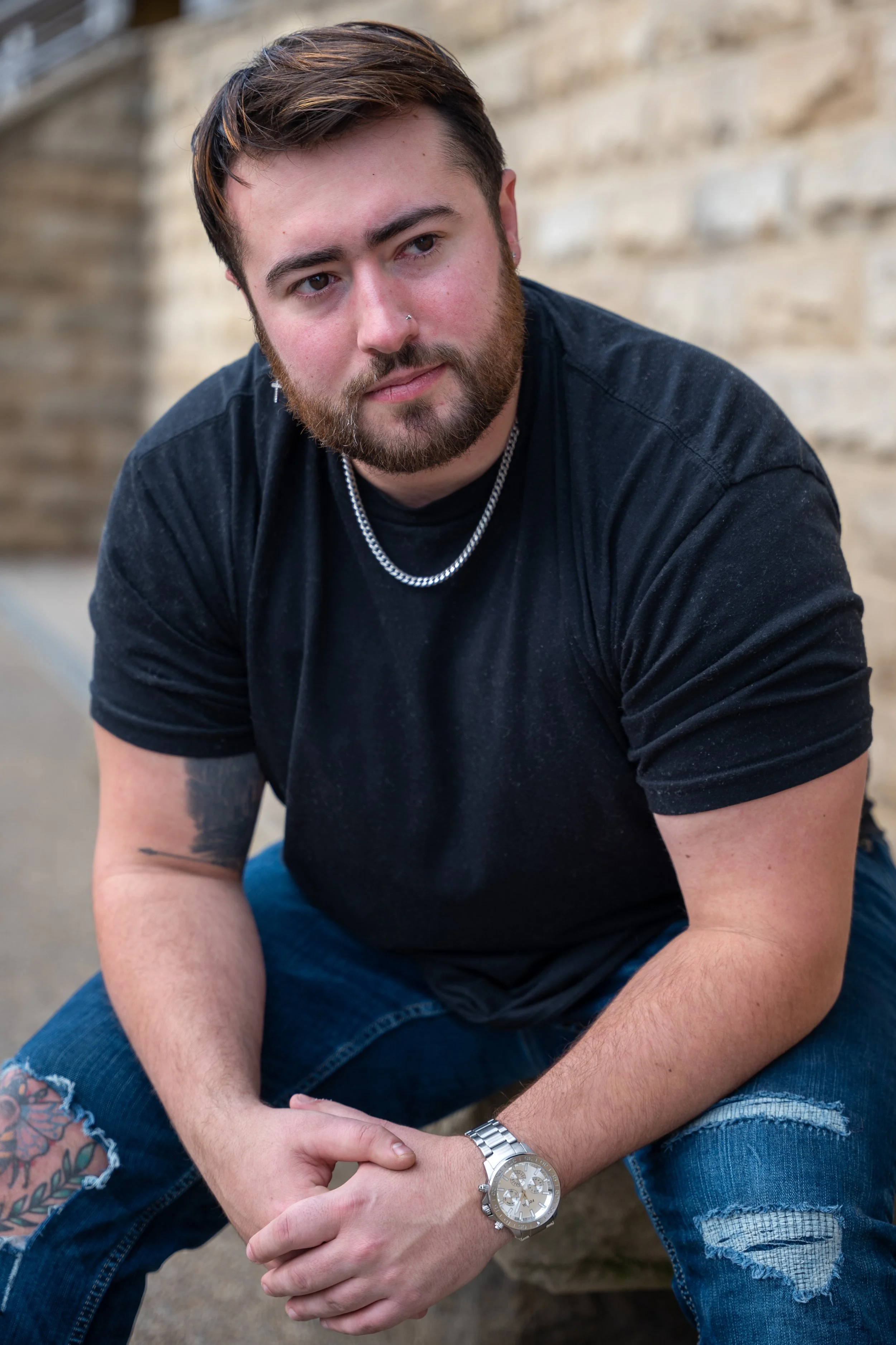 A young man with a beard and mustache, dressed in a black t-shirt and ripped jeans, sitting outdoors near a brick wall, wearing a silver watch, a chain necklace, and small hoop earrings.