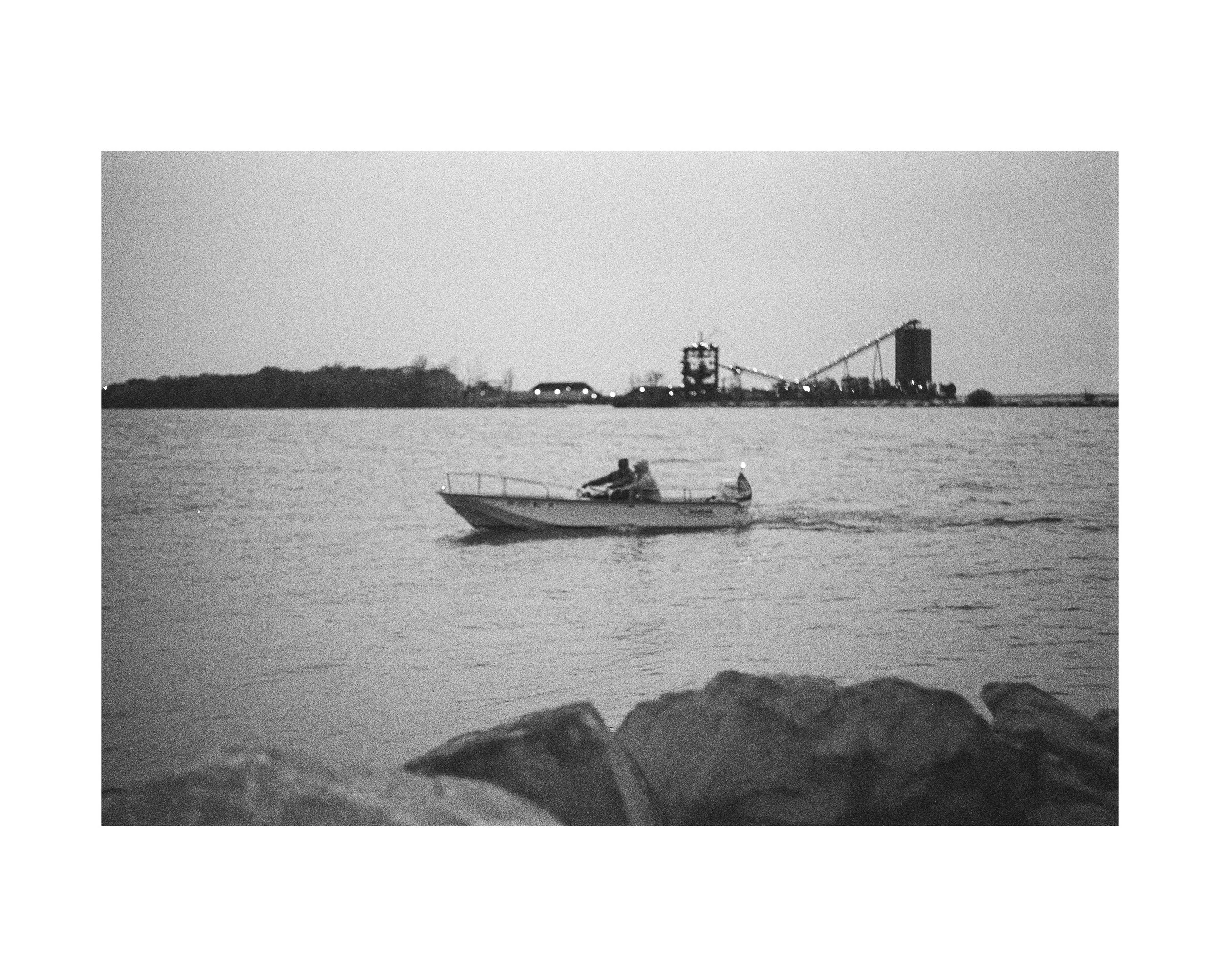 A boat with people sailing on a body of water, with rocks in the foreground and an industrial area or factory with tall structures and smoke in the background, in black and white.