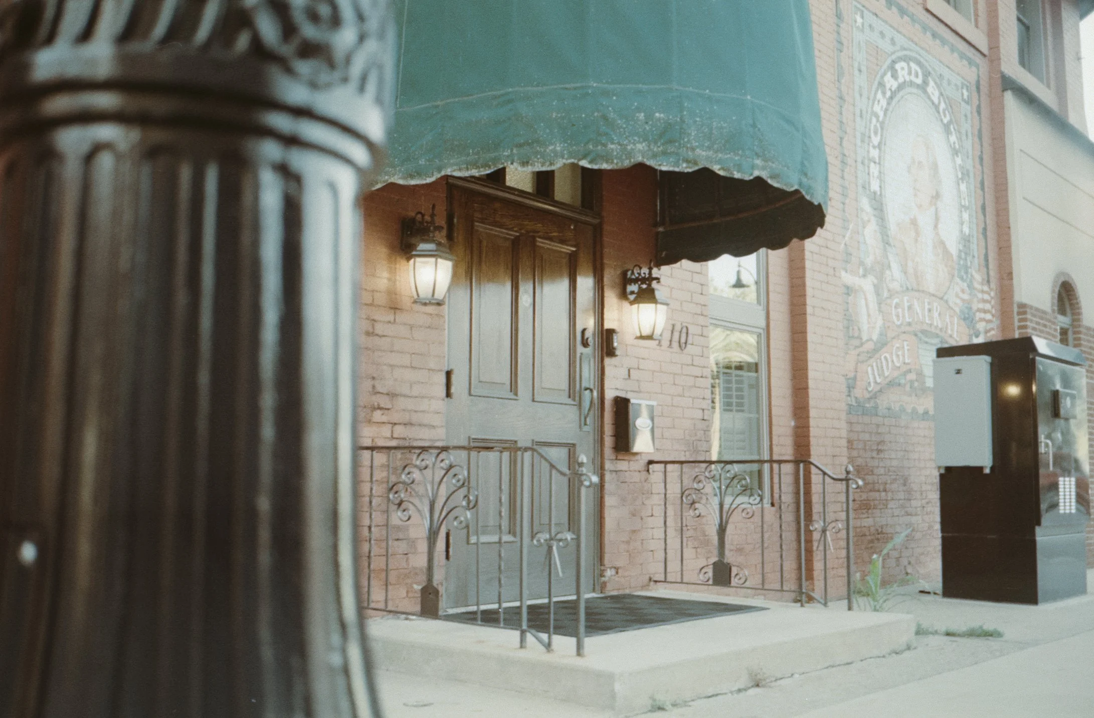 The entrance of a building with brick walls, a wooden door, two wall-mounted lanterns, a green awning, and a metal railing leading up to the door. There is also a mosaic sign on the wall that reads 'Board of County General Judge.'