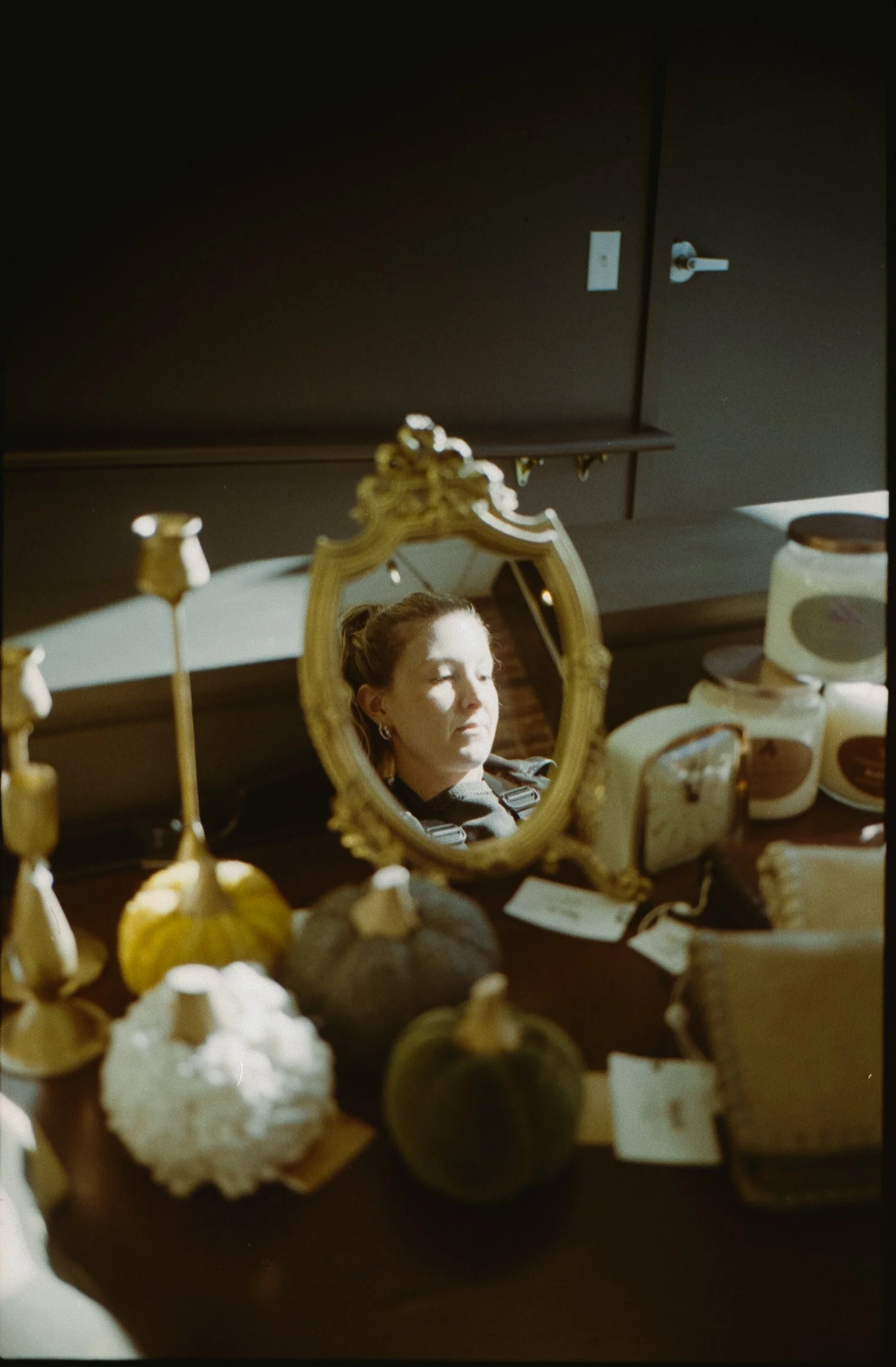 A woman is seen in her reflection in a small ornate mirror on a table, surrounded by pumpkins, candles, and skincare or cosmetic products.