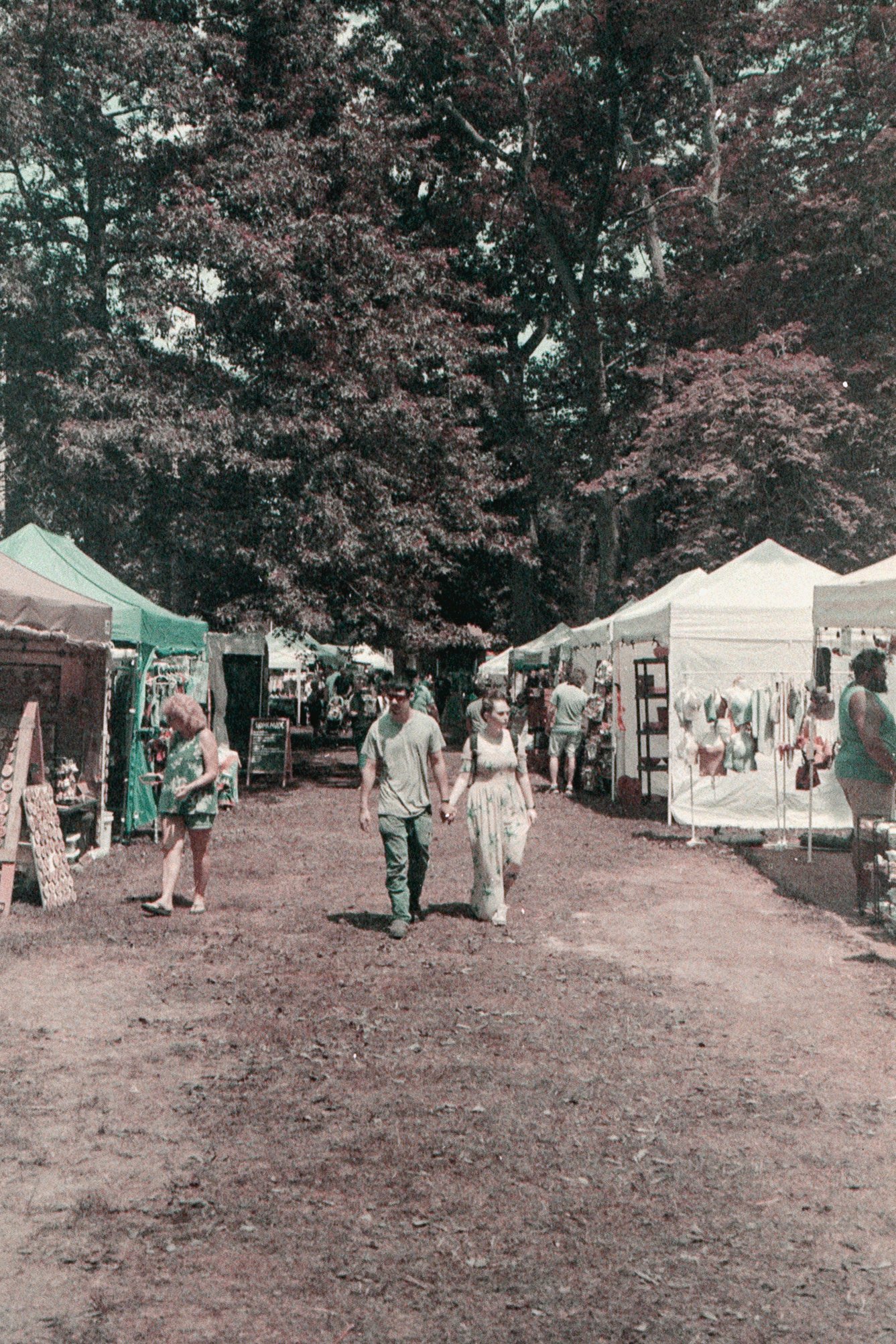 People walking in a marketplace or fair with white tents and trees in the background.