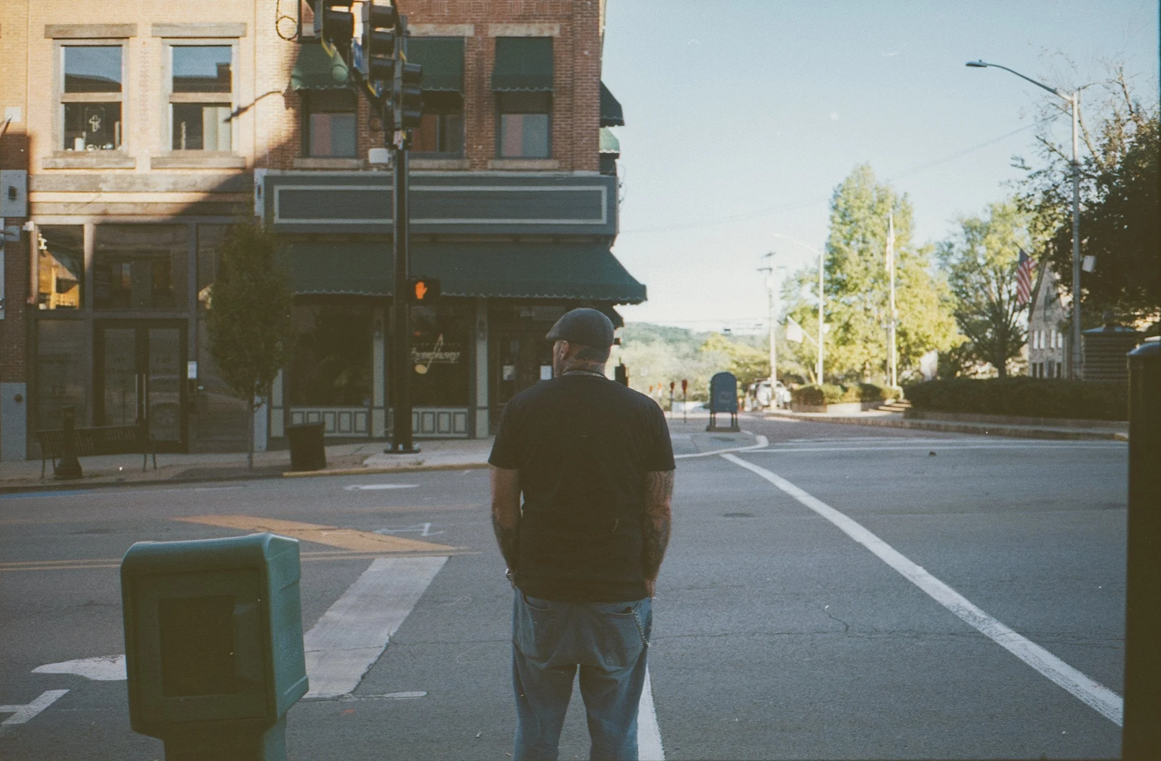 A man with tattoos on his arms, wearing a black T-shirt, gray sweatpants, and a beanie hat, stands at a crosswalk on a quiet street. There are buildings, a bench, and trees in the background, with a clear sky.