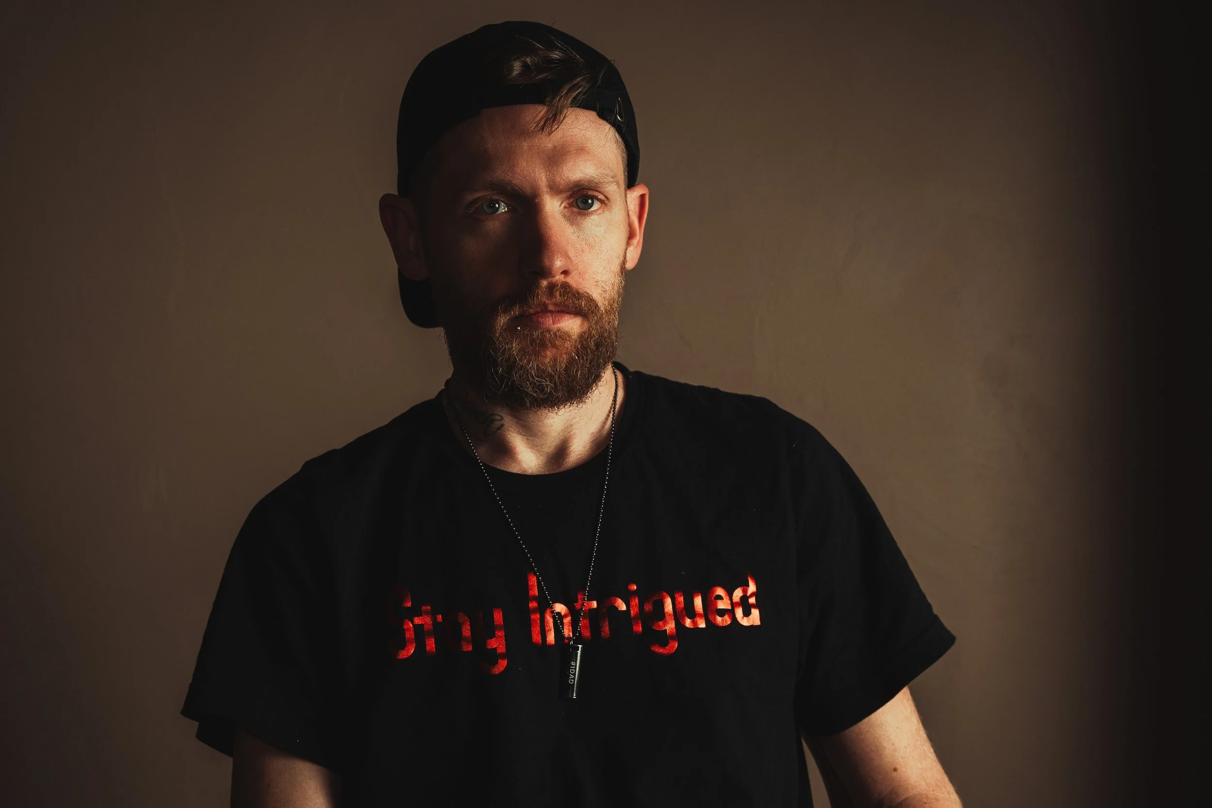A young man with a beard, wearing a black backwards cap and a black T-shirt with red text, stands against a dark beige background. The lighting highlights one side of his face.