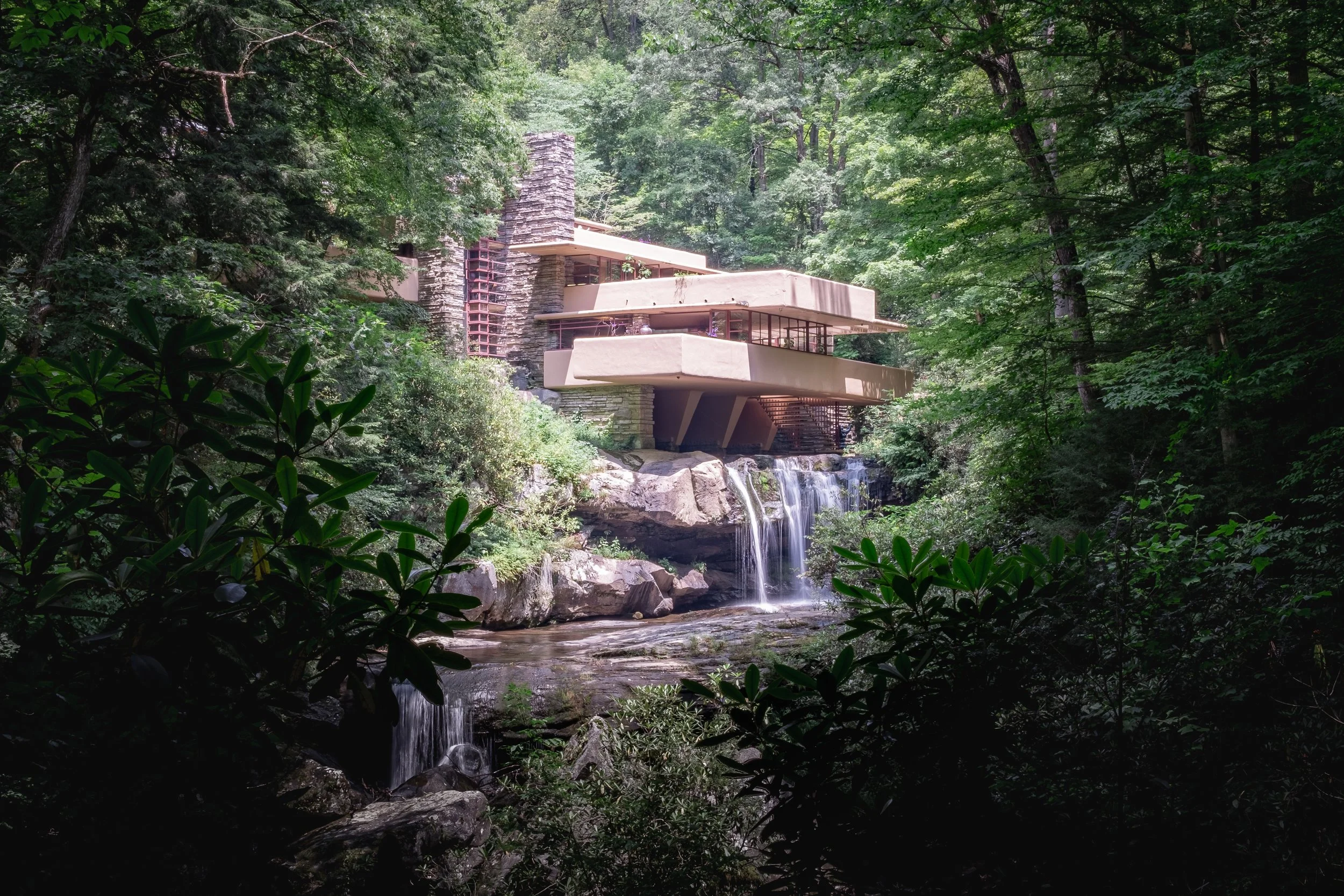 Falling water surrounded by lush green trees with a modern house built on rocks over the waterfall.