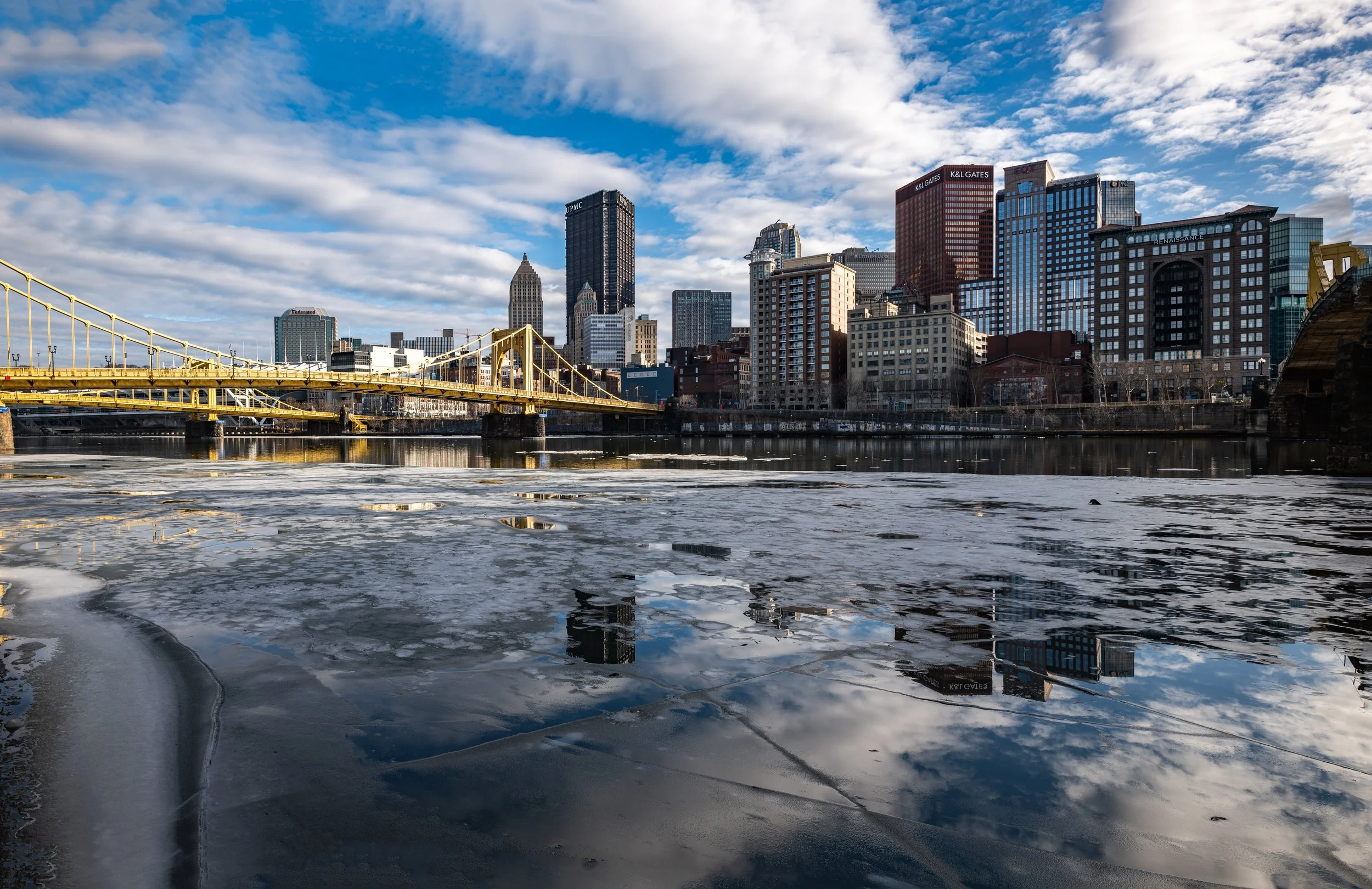 City skyline with skyscrapers and bridge over partially frozen river under a partly cloudy sky.