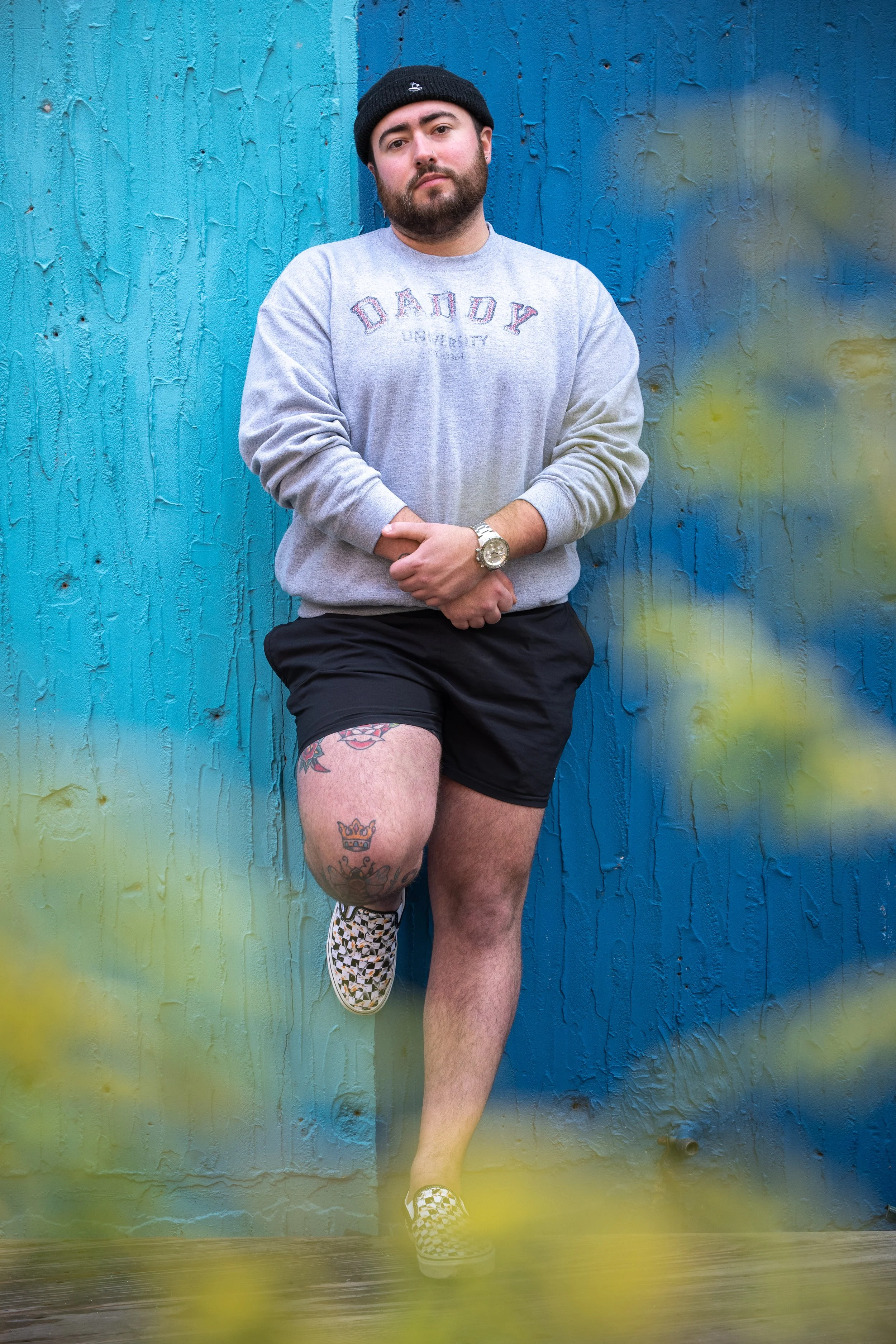 Portrait shot of a man wearing a black beanie, gray sweatshirt with 'DADDY' written on it, black shorts, and checkered slip-on shoes, standing against a blue wooden wall, with one leg raised and hands clasped.