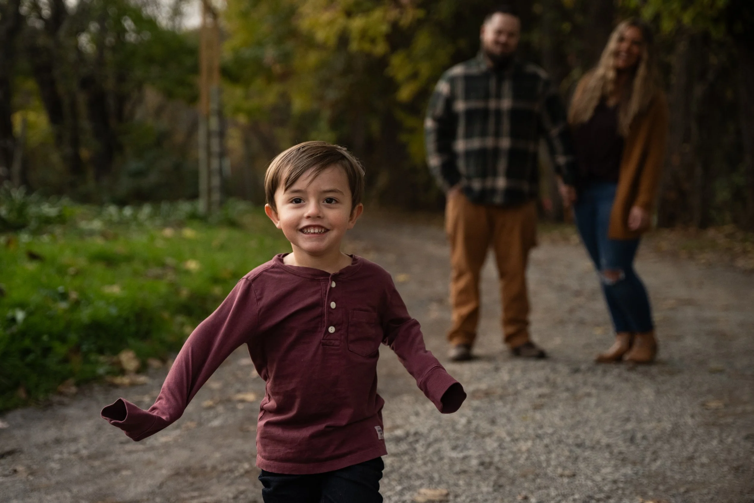 Family portrait of a young boy running on a dirt path in a forest, smiling at the camera, with a man and woman holding hands in the background.