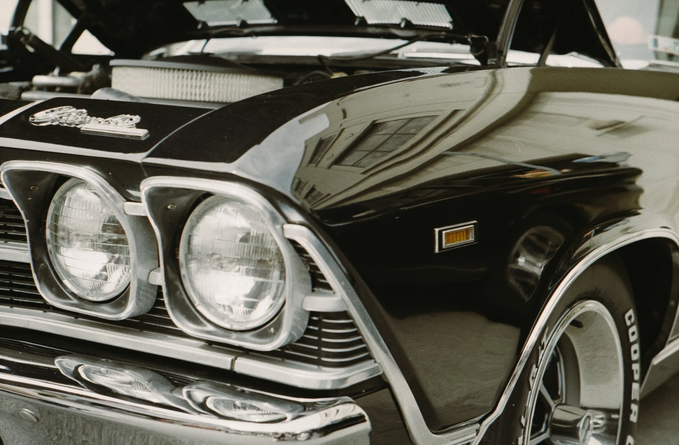 A close-up of the front of a vintage black muscle car, showing dual round headlights, chrome accents, a prominent hood ornament, and a black exterior with chrome details.