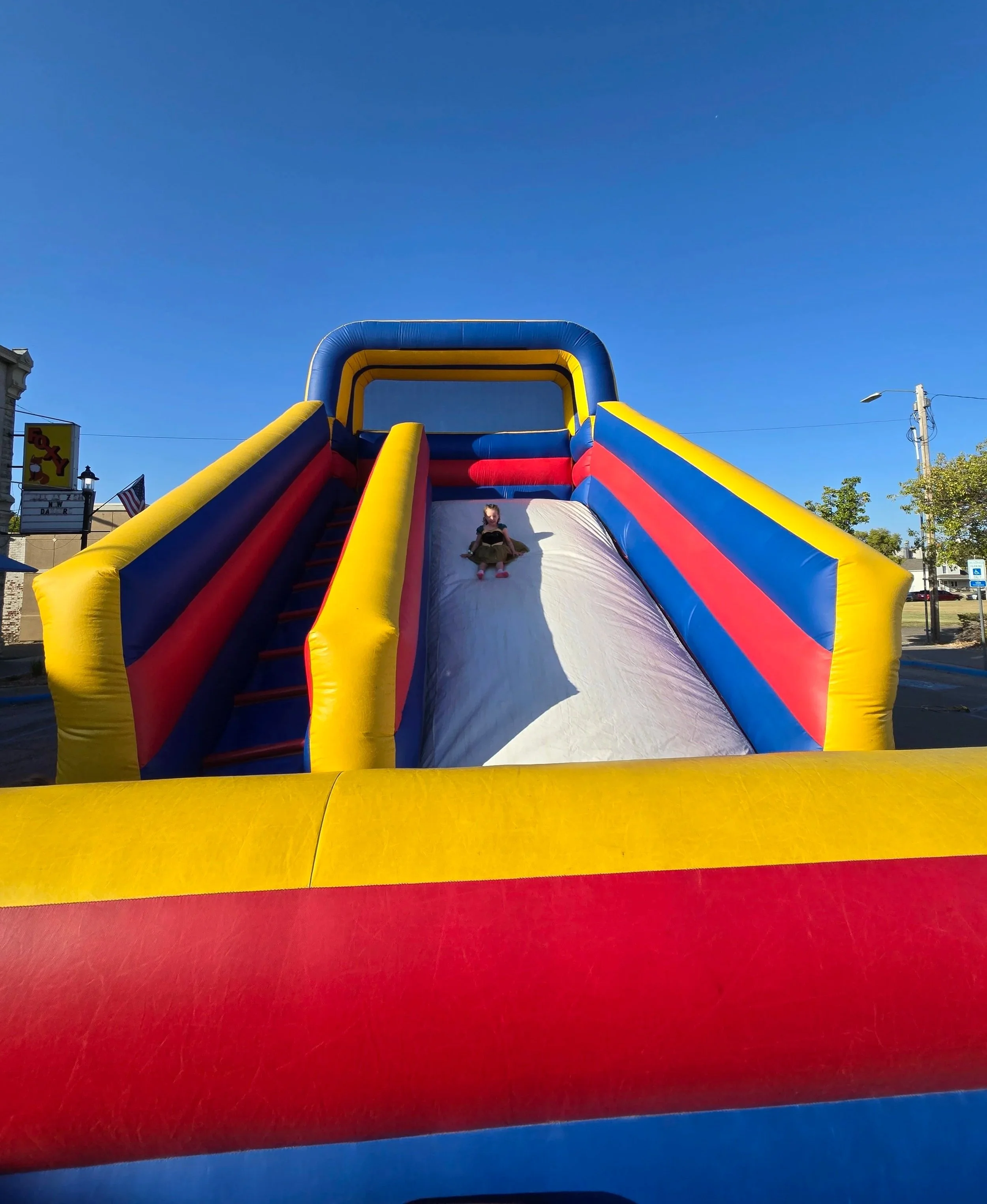 Child sliding down an inflatable colorful bounce slide, blue sky background, part of a fun outdoor event.
