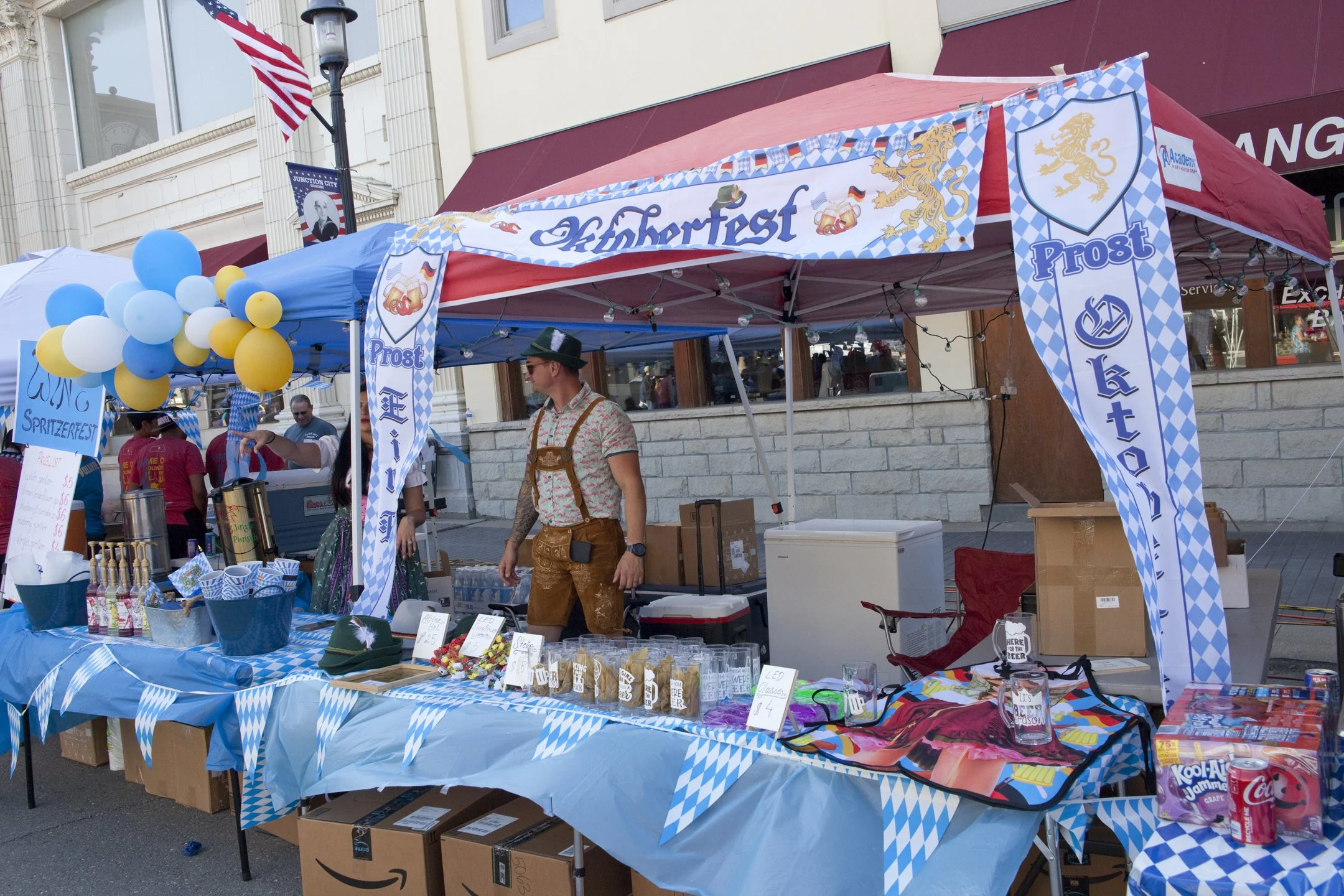 Outdoor Oktoberfest booth with blue and white decorations, including balloons and banners, offering drinks, merchandise, and memorabilia.