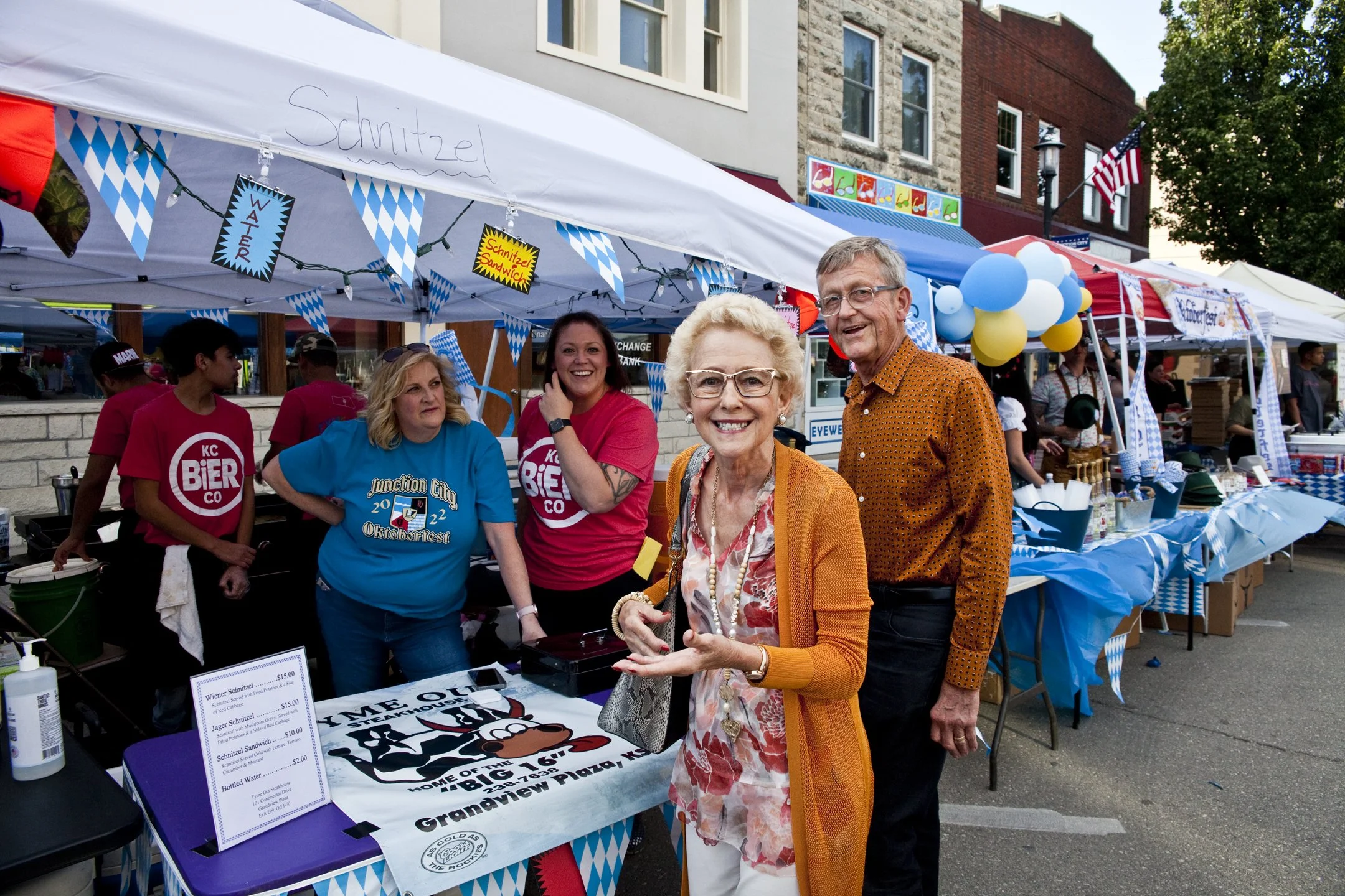 Happy elderly woman in orange cardigan and glasses talking, standing next to an elderly man in orange polka dot shirt, at a street fair with blue and white tents, balloons, and people in the background.