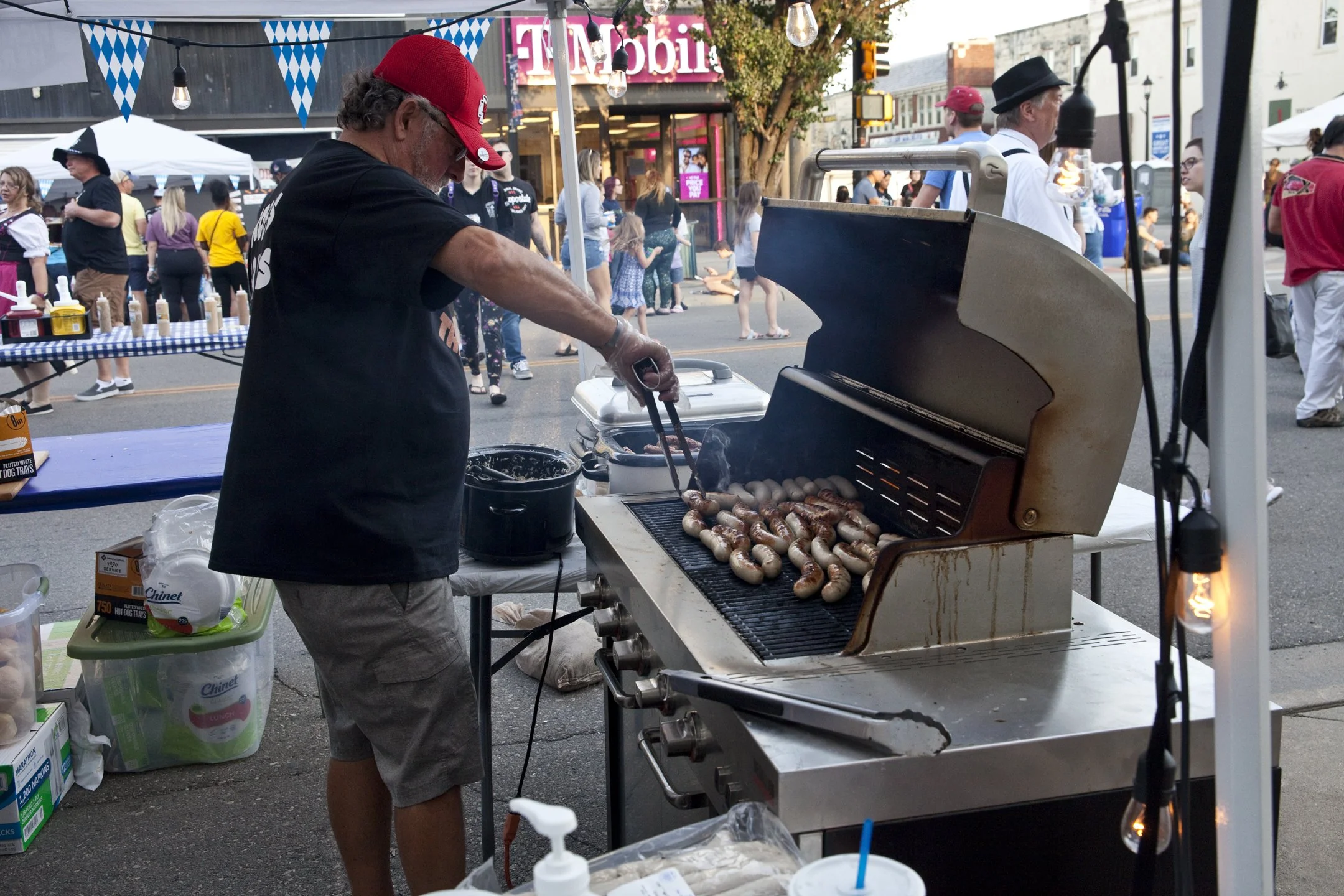 Man grilling sausages and hot dogs at an outdoor street festival, with many people walking and shopping in the background.