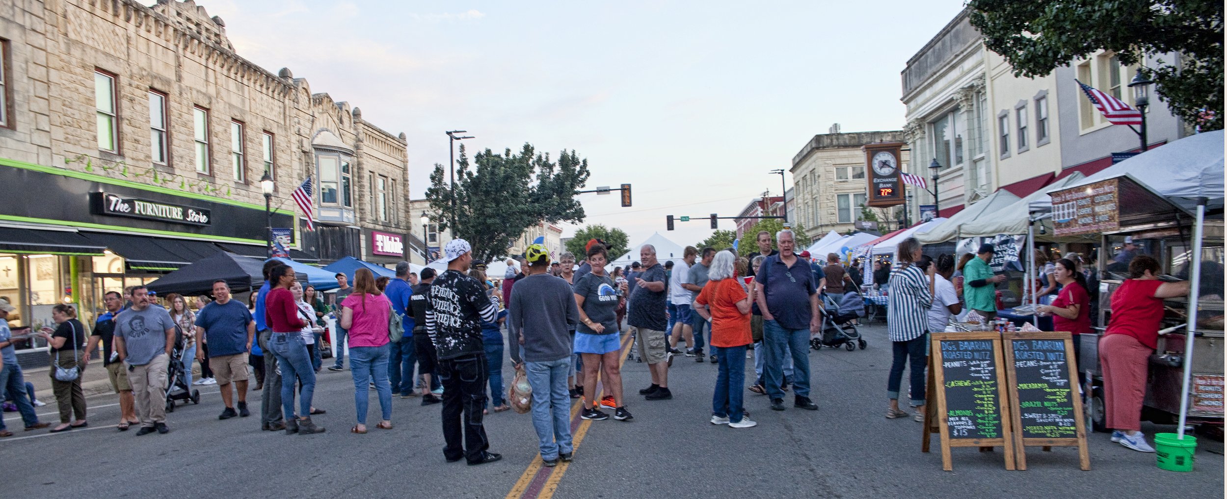 Crowd of people walking and shopping at an outdoor street market with tents and food stalls, buildings on either side, and a traffic light in the background.