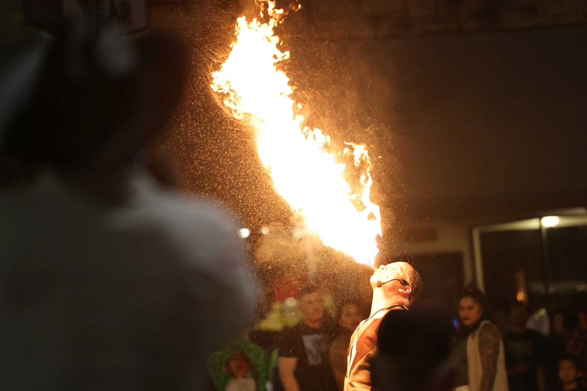 Performer in a fire show breathing a large flame while an audience watches in the background.
