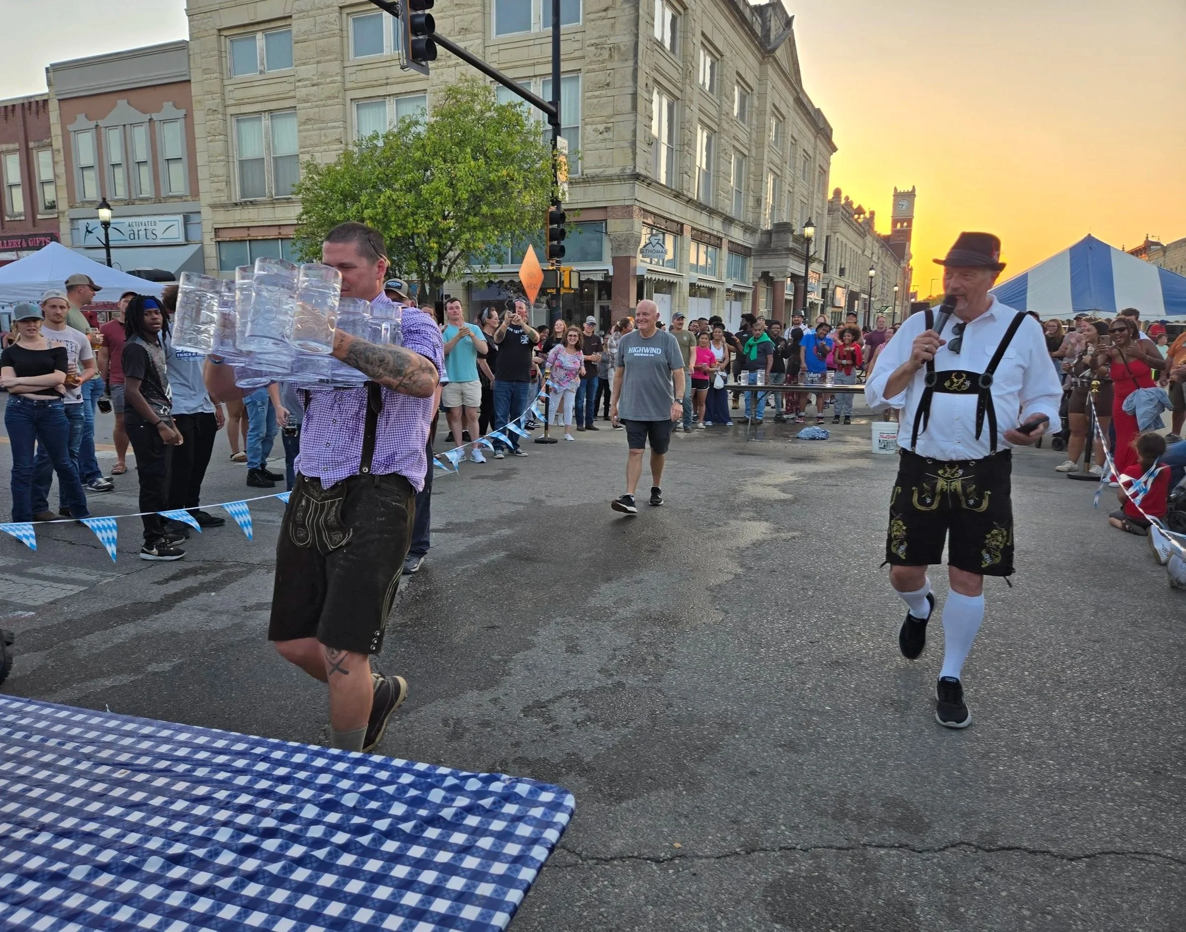 A man in traditional Bavarian clothing holding a microphone, and another man carrying multiple beer mugs on a tray at an outdoor festival during sunset, with a crowd of people watching.
