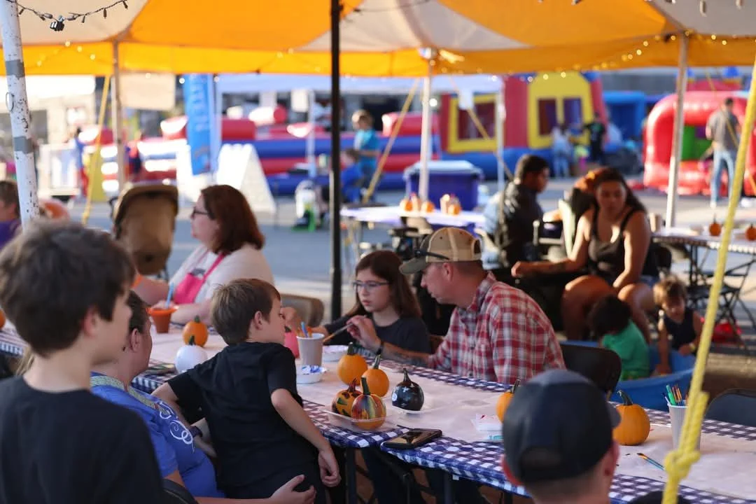 A group of children and adults sitting under a yellow canopy at a fall-themed outdoor event, with pumpkins on a checkered tablecloth and other amusement rides in the background.