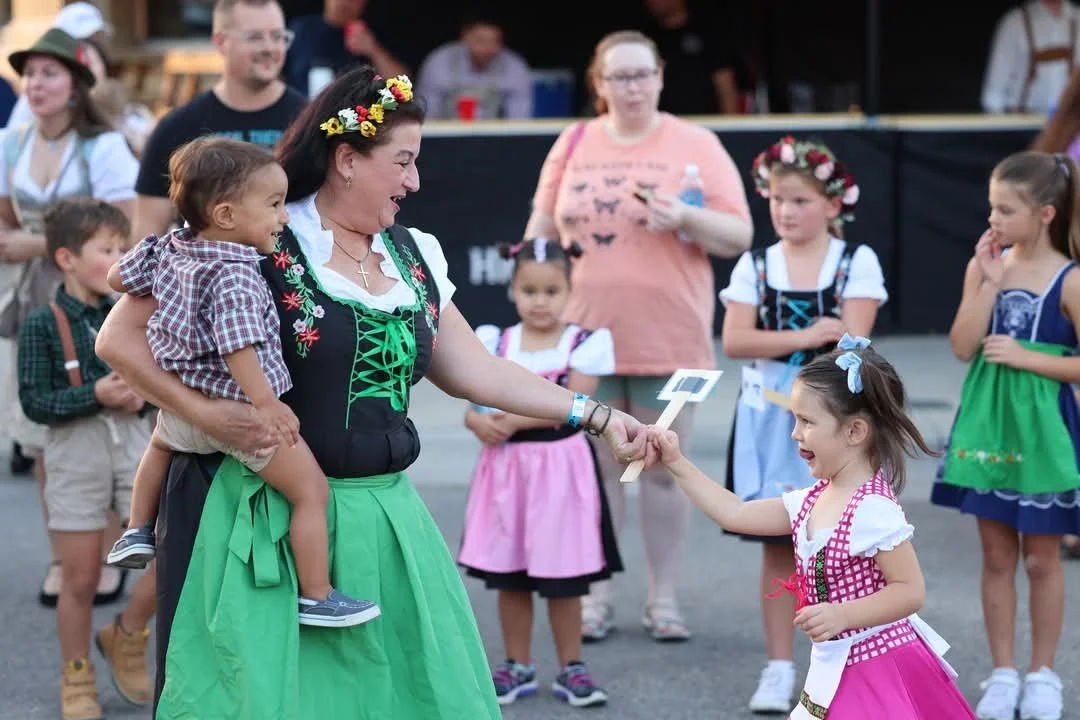 A woman in traditional German dress holding a young boy, shaking hands with a young girl in similar dress at an outdoor festival with other children and adults in the background.