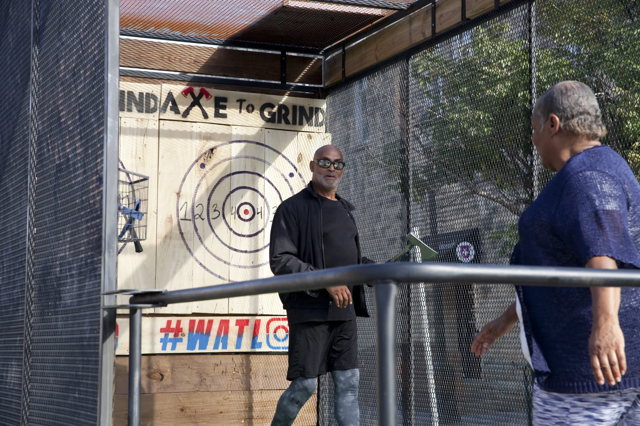 Two men playing axe throwing games at an outdoor range with a target and safety cage, one man holding an axe and the other observing, under a wooden and metal roof with trees in the background.