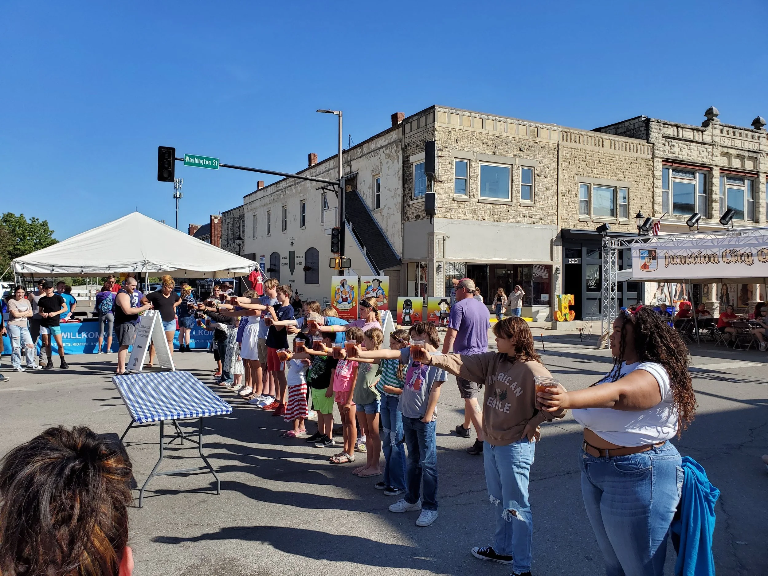 People participating in a parade or festival, standing in a line with their arms extended forward, holding drinks, in a city street with buildings and a tent in the background.