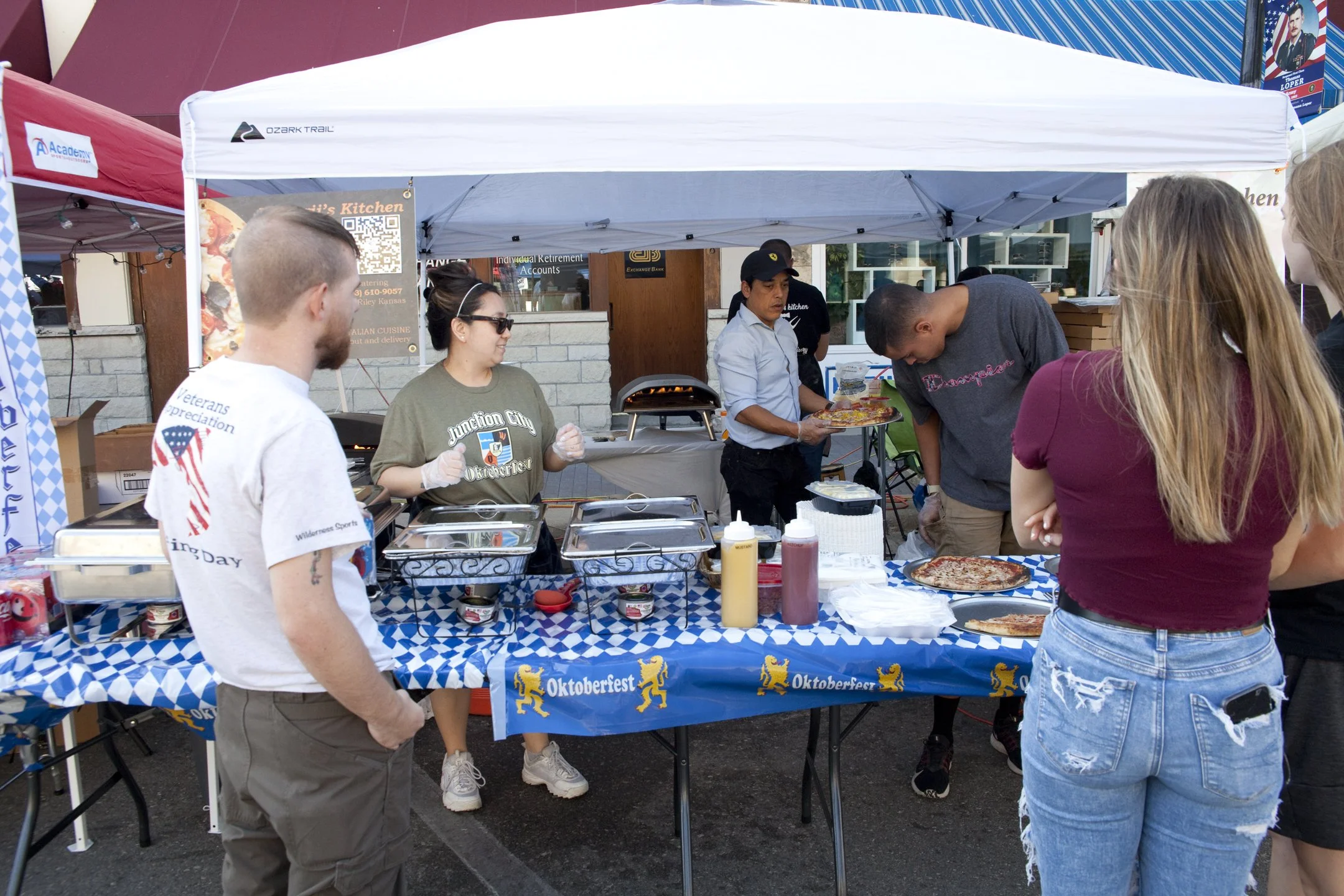 People at an Oktoberfest food stall with pizza, sauces, and condiments, serving and ordering food under a white canopy.