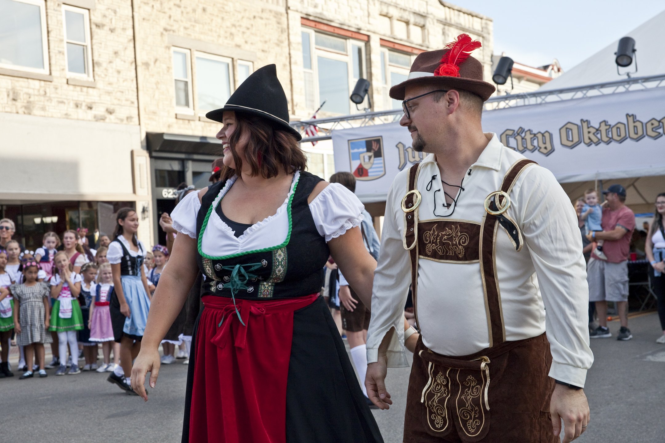 A man and woman dressed in traditional German clothing walking together at a cultural event, with a crowd and festival banner in the background.