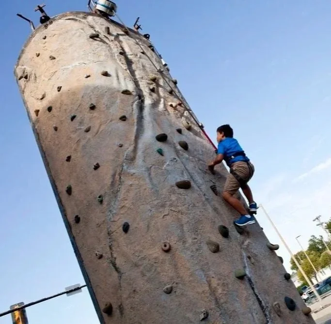 A young boy climbing an outdoor rock wall under a clear blue sky.