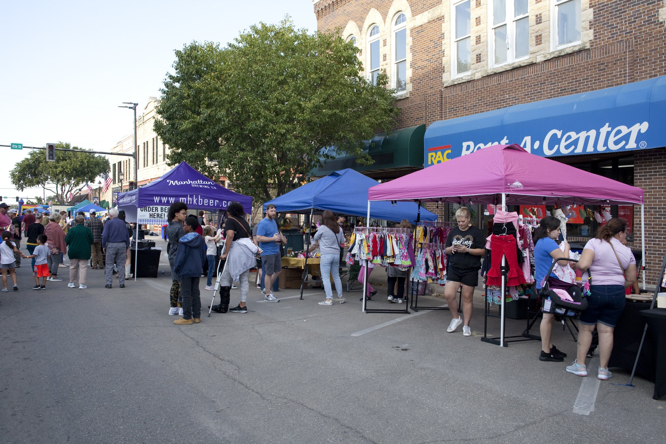 Outdoor street market with colorful tents and vendors, people shopping and walking on sidewalk, brick buildings, tree, and traffic light in background.