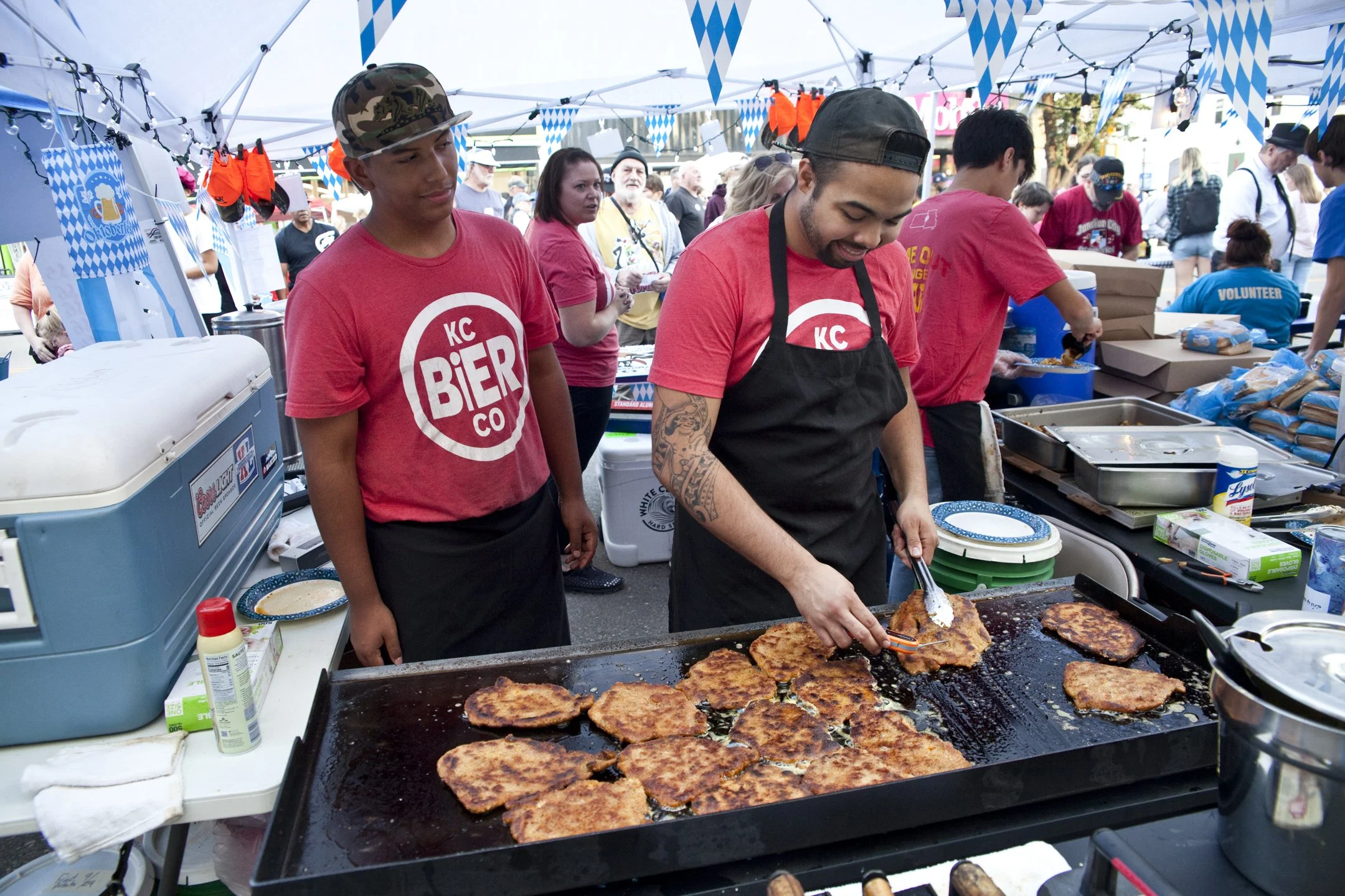 Two men cooking large pieces of meat on a flat-top grill at an outdoor event decorated with blue and white balloons and flags. One man is wearing a red KC Bier Co t-shirt and cap, the other is wearing a red t-shirt and black apron, with tattoos on hi