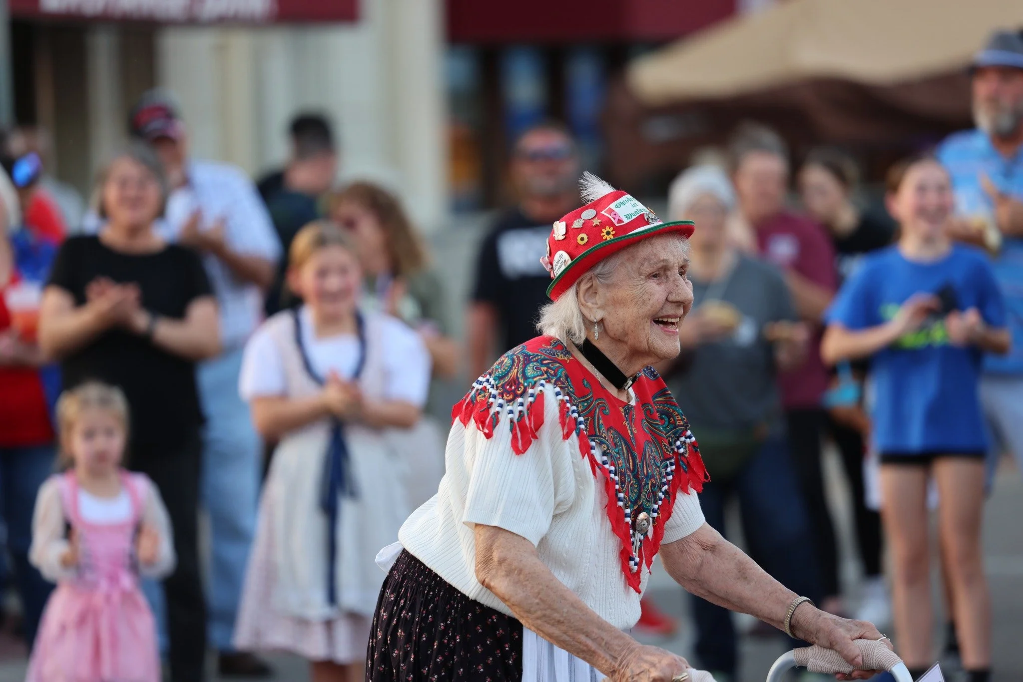 An elderly woman dressed in a traditional folk costume, wearing a colorful hat and blouse, smiling while holding a cane at a public outdoor event with a crowd of people in the background.
