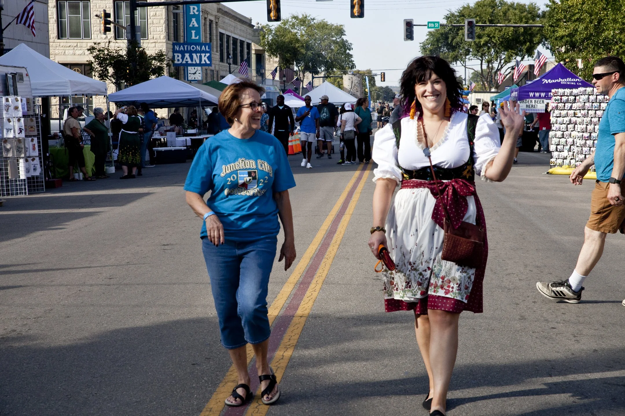 People walking at an outdoor street festival with tents and booths, some flags, and a building with a sign that reads 'NATIONAL BANK' in the background.