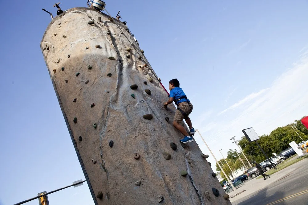 Child wearing a blue shirt and brown shorts climbing an outdoor rock wall with handholds, under a clear blue sky, in a paved area with trees and parked cars nearby.
