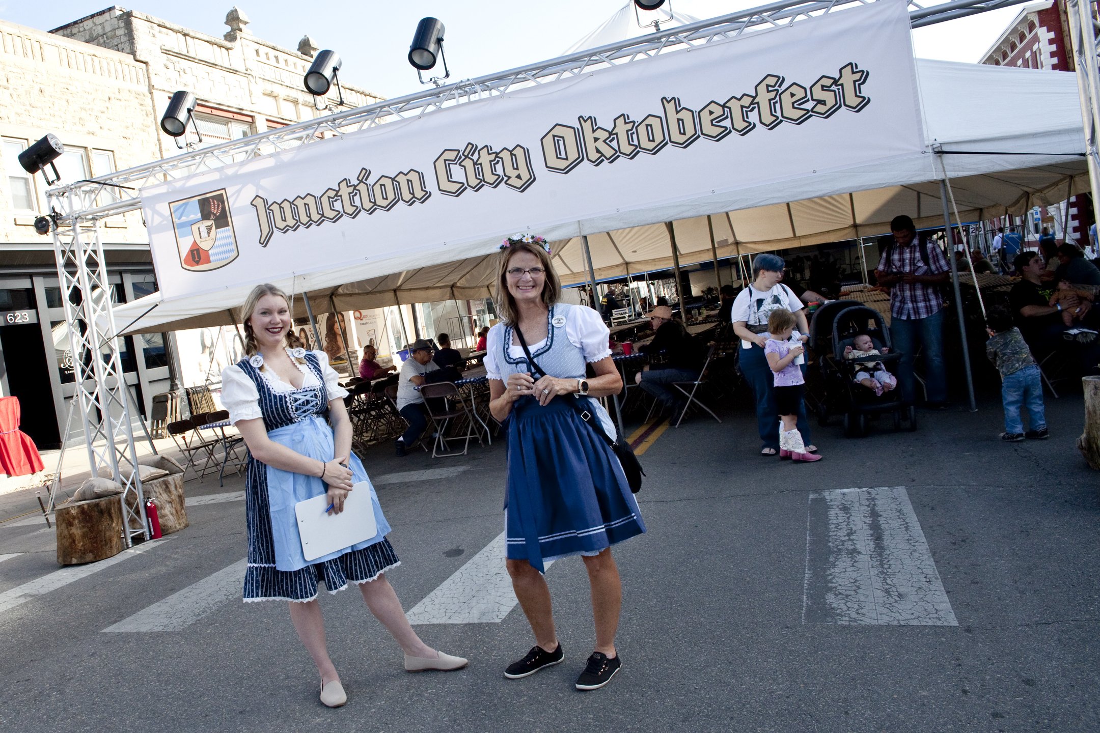 Two women dressed in traditional German dirndl dresses standing in front of a tent with a sign that reads 'Junction City Oktoberfest'. The women are smiling, with one holding a clipboard, and there are people sitting and walking in the background at 
