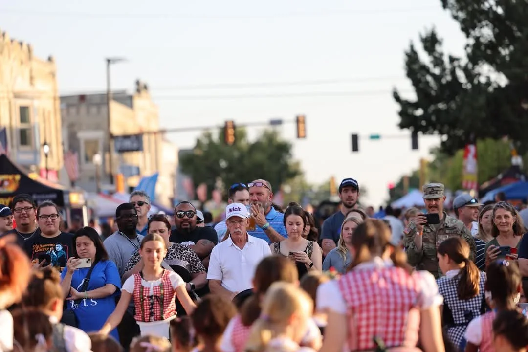 Crowd of people gathered outdoors at a festival or event, some taking photos or videos, others watching a performance, with tents and buildings in the background.