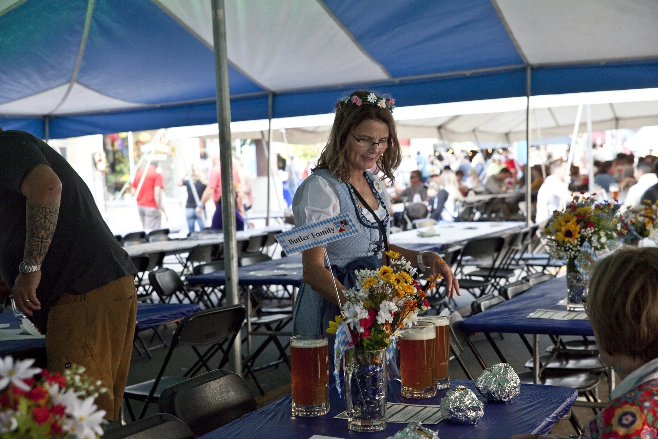 A woman dressed in traditional Bavarian attire standing at a table with three large beers and floral decorations during an outdoor festival, with many people seated and walking in the background.