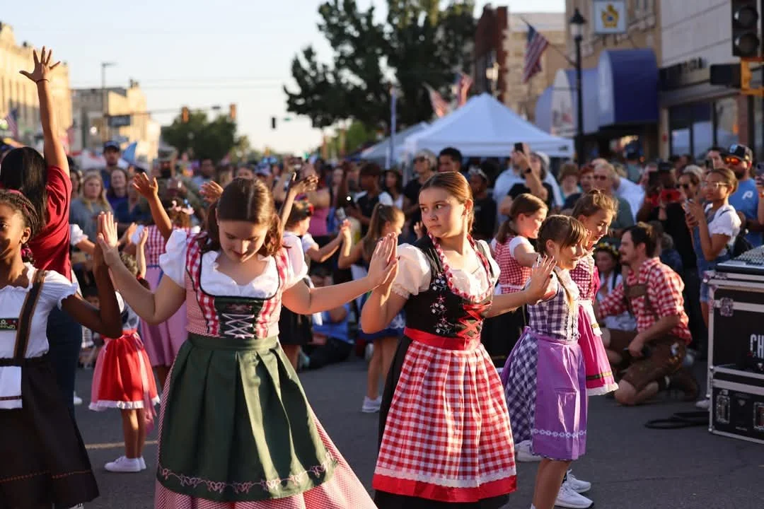 Young girls in traditional dresses dancing in a street festival with an audience watching.