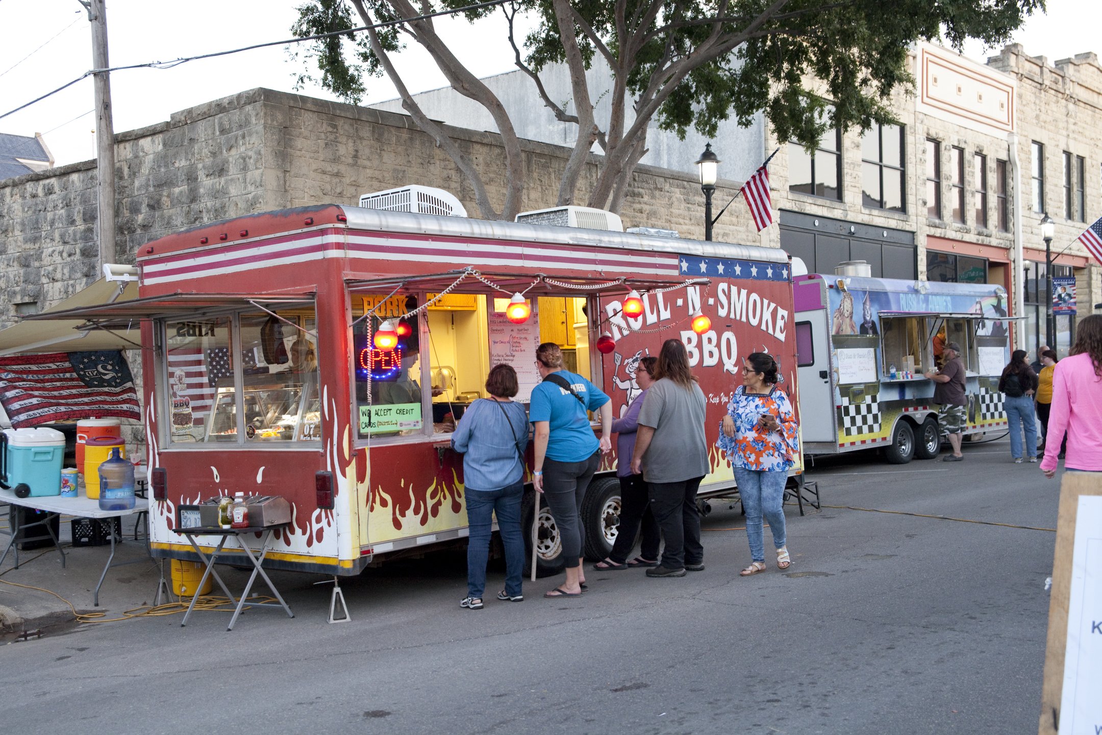 People ordering food at a red barbecue food truck with flame graphics, sellin barbecue and smoked food, parked on a city street with other food trucks and pedestrians.
