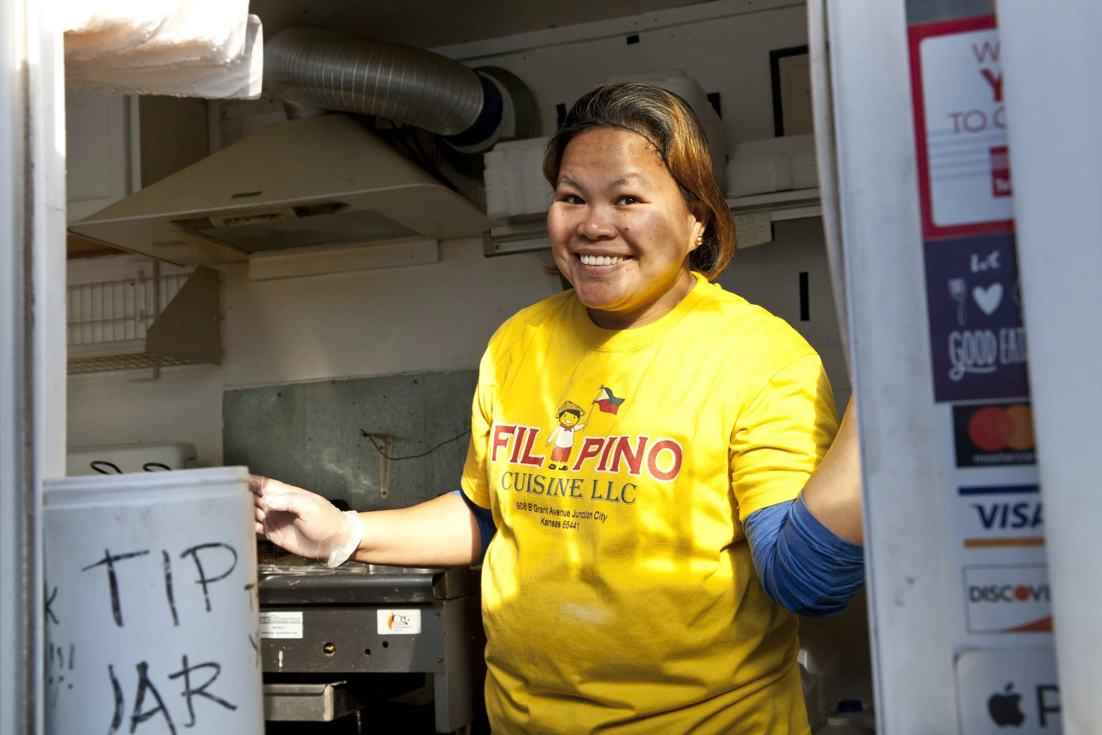 A smiling woman wearing a yellow T-shirt with 'FILIPINO CUISINE LLC' written on it, standing in a food truck or small kitchen, reaching for a tip jar.