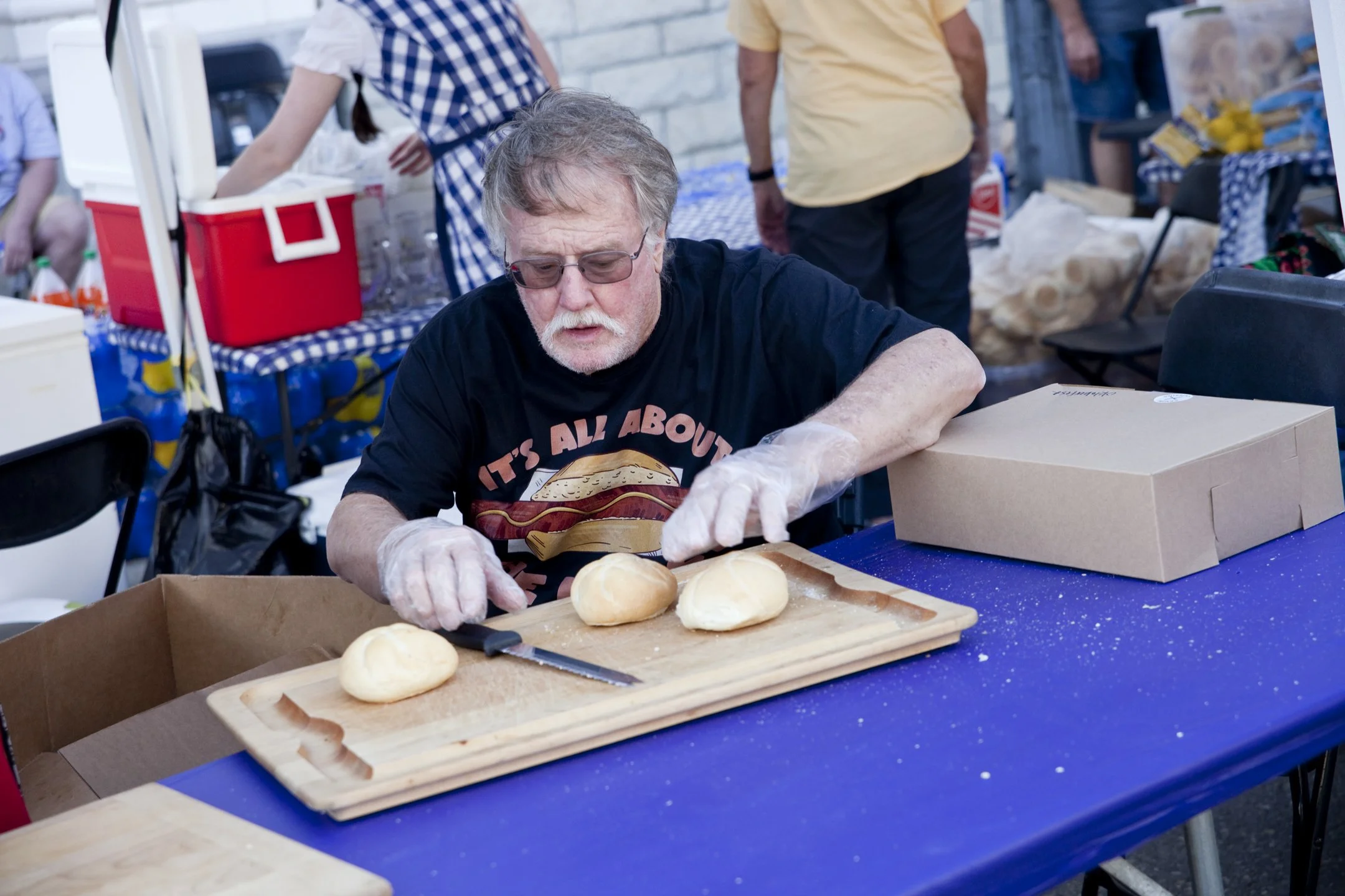 An elderly man with glasses and a beard wearing a black T-shirt, preparing buns at an outdoor event.