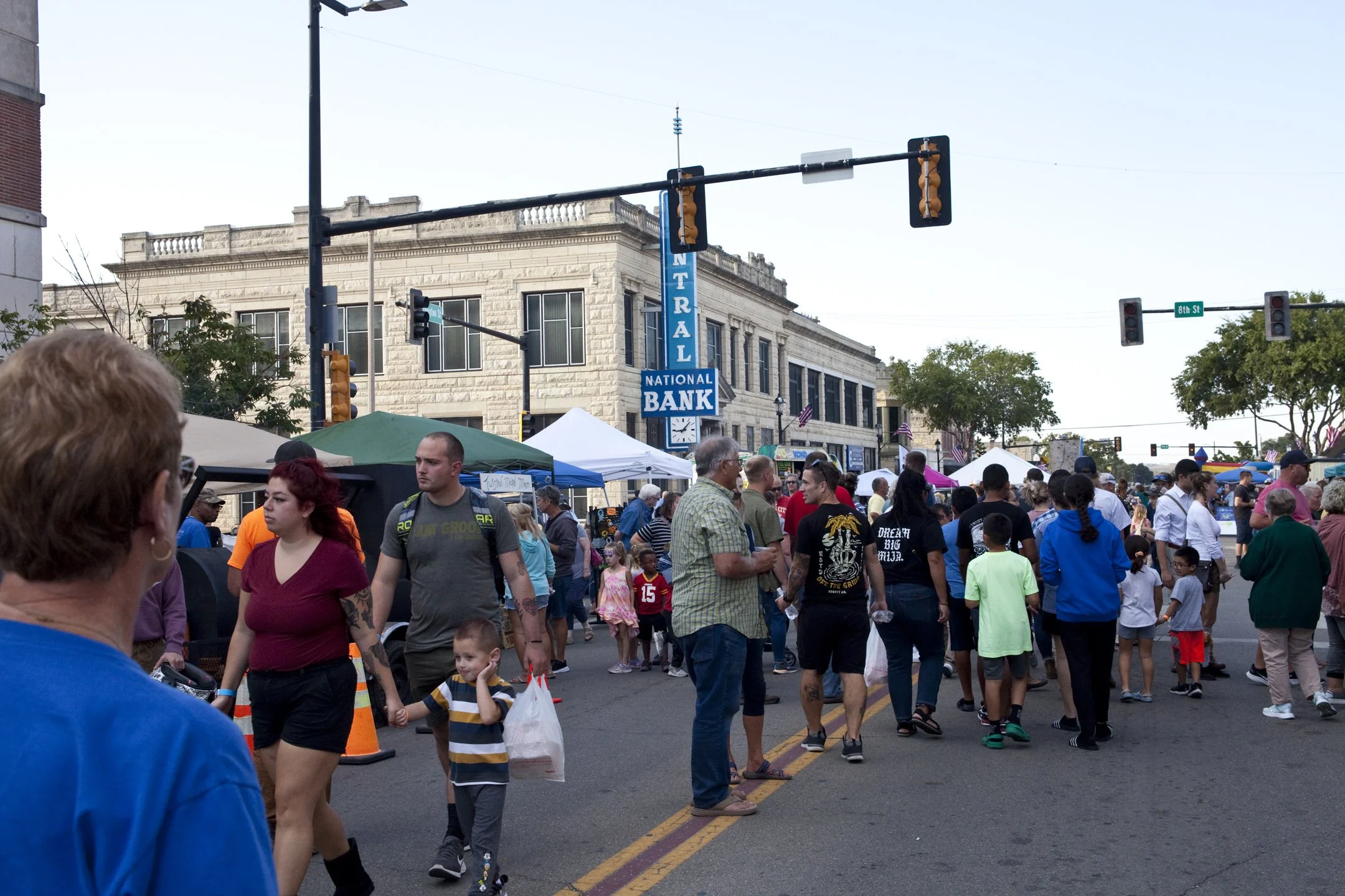 Crowd of people at an outdoor street market in front of a building with a sign that reads 'NATIONAL BANK'. Various tents and stalls line the street, with trees and traffic lights overhead.