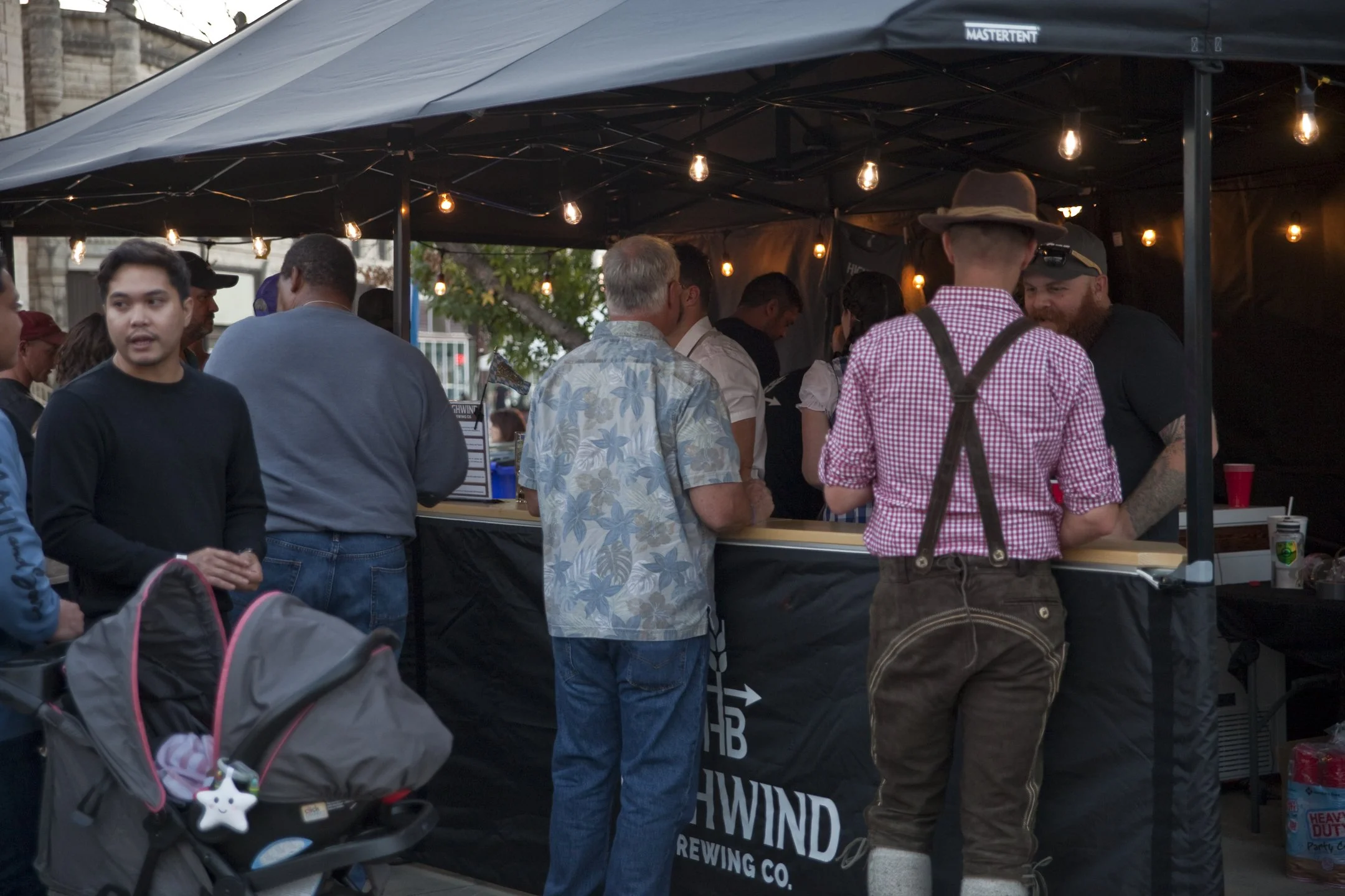 People standing at a food or drink stall at an outdoor event with string lights hanging above. One person is dressed in traditional German attire with suspenders and a hat.