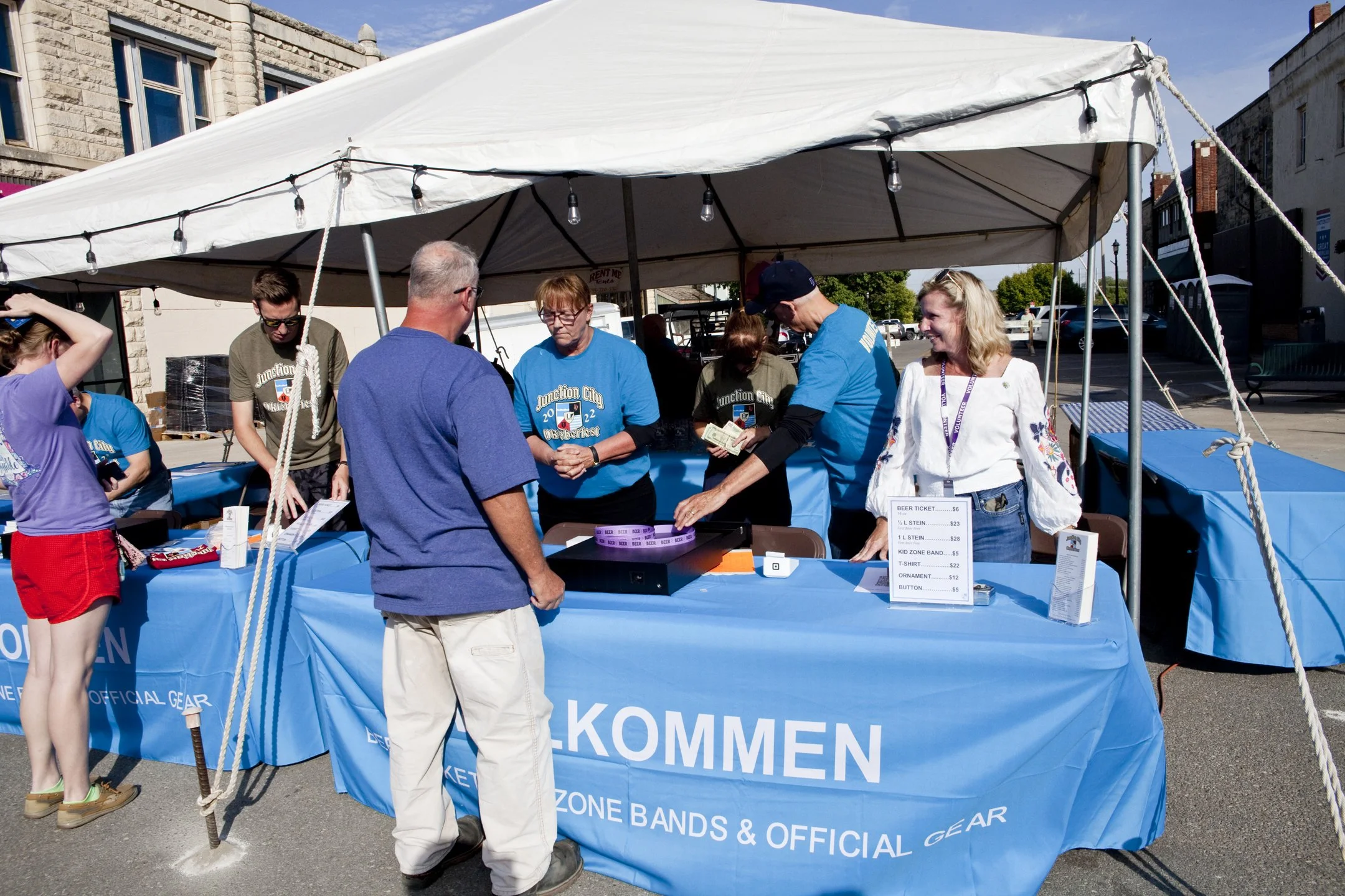 People at an outdoor event under a white tent, engaging in a ticket-selling activity with a blue tablecloth that says 'KOMMEN' and involves bands and official gear.