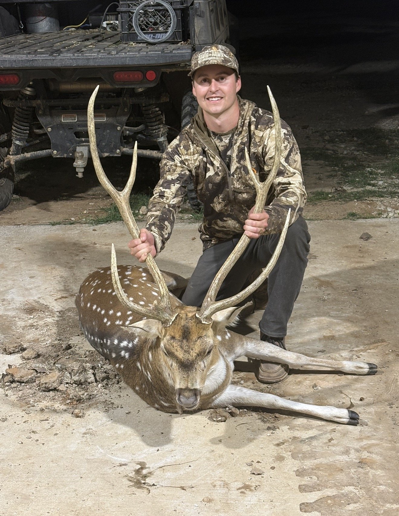 A man in camouflage attire kneels next to a fallen deer, holding its antlers. The deer has white spots on its brown body, and the scene appears to be outdoors at night.