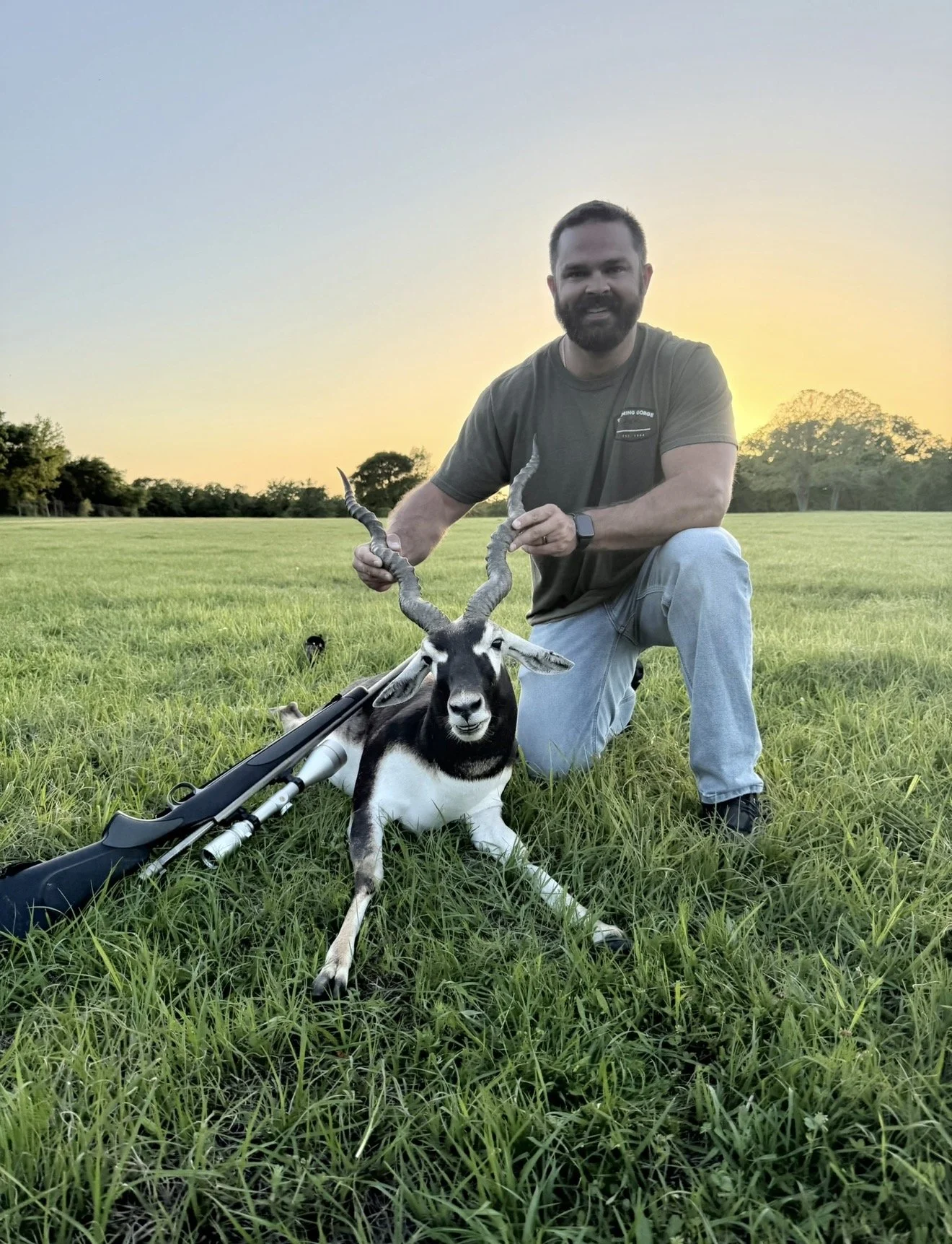 A man kneeling on grass in a field at sunset, holding the horns of a black and white antelope with long twisted horns, with a rifle lying on the ground beside it.