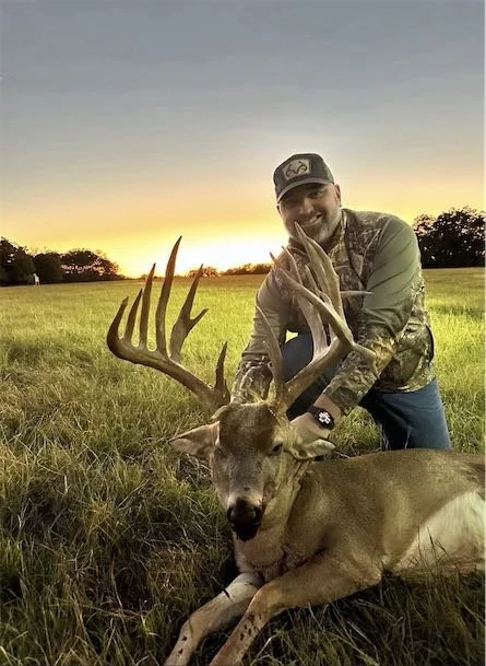 A man in camouflage clothing and a cap smiling while kneeling behind a large, dead deer with impressive antlers in a grassy field at sunset.