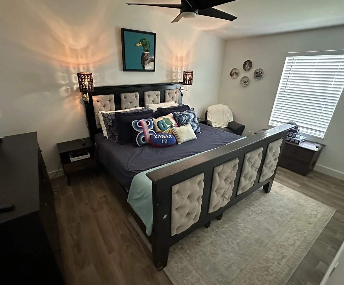 A bedroom with a black wooden bed frame with beige tufted upholstery on the headboard and footboard. The bed has multiple pillows, including one that says 'XANAX 1.0' and others with colorful designs. There are two black nightstands on either side of