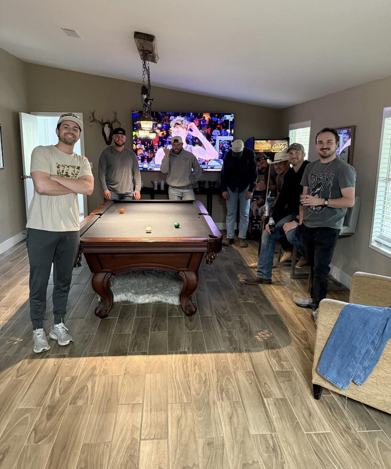 A group of eight men gathered in a living room for a casual get-together, standing around a pool table with a television in the background displaying a sports event.