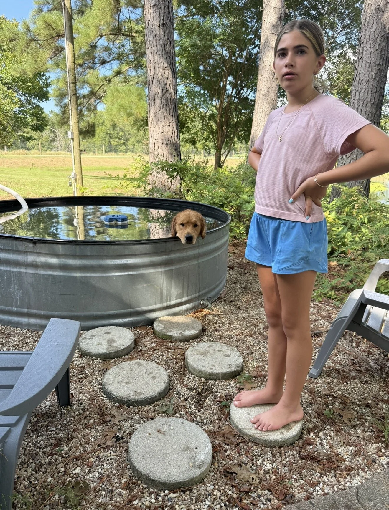 A young woman standing outdoors on a rock stepping stone in front of a small above-ground pool with a dog resting its head on the edge. The woman is wearing a pink T-shirt and blue shorts, with her hands on her hips. The background shows trees, green