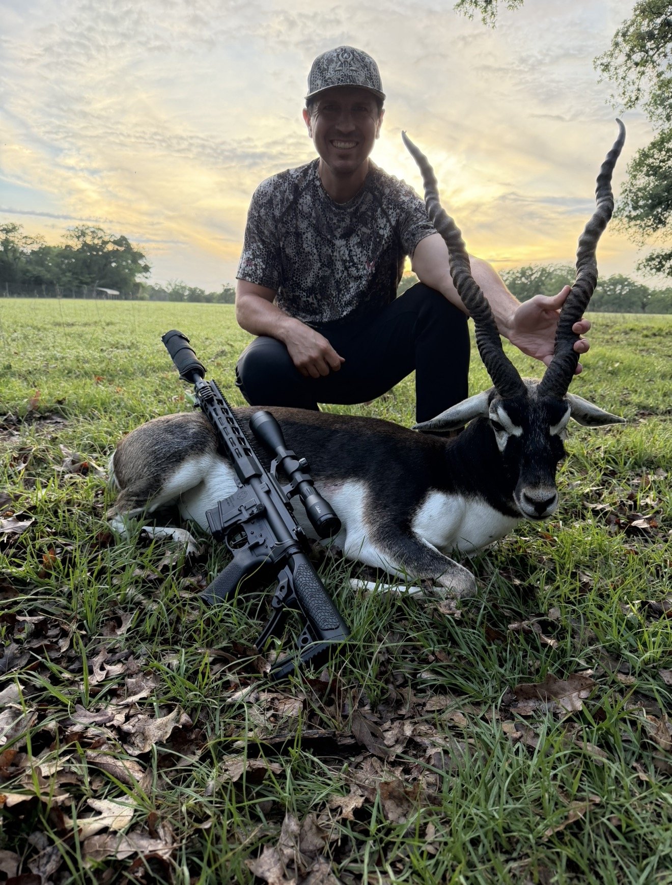 A man kneeling outdoors during sunset, smiling, holding a goat with twisted horns, with a rifle resting on the ground nearby.